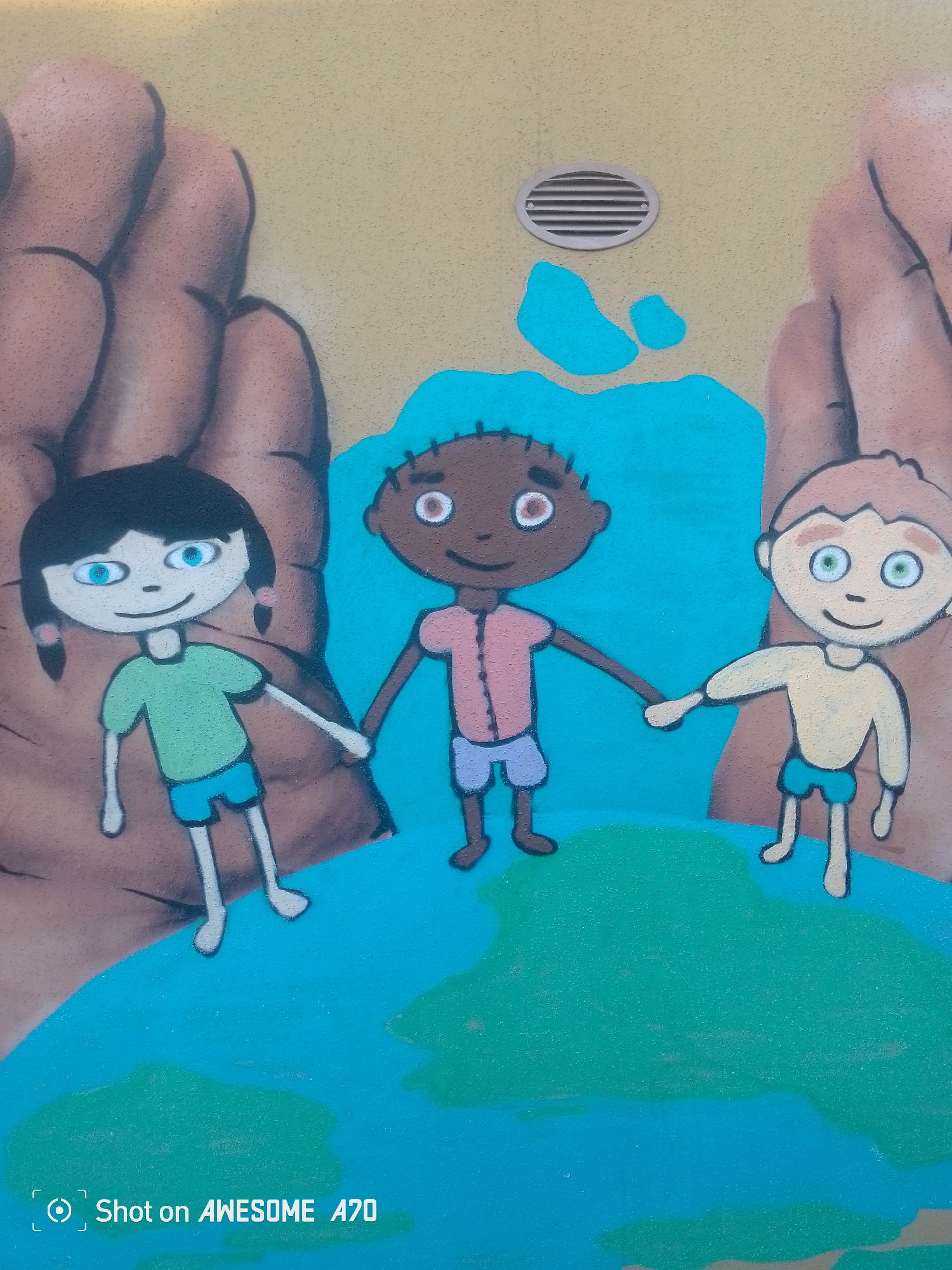 This is a close-up, eye-level shot of a mural painted on a textured, light tan wall. The mural depicts three cartoon-style children holding hands and standing on a representation of the Earth. The children are diverse in appearance: one has dark hair and pigtails with blue eyes, another has dark skin and brown eyes, and the third has light skin and green eyes. They are all wearing simple, brightly colored clothing.
The background features two large, stylized hands, one on each side, seemingly cradling the children and the Earth. A section of bright blue paint, possibly representing water or sky, is behind the children, and to the left of that, a small, oval-shaped ventilation grate is visible on the wall. The Earth is depicted with green landmasses and blue water.
The overall impression is one of unity and global connection, conveyed through the children's clasped hands and their position on the Earth. The mural appears to be outdoors, possibly on a public building, given the presence of the ventilation grate. There are no obvious cues about the time of day or weather.
In the bottom left corner of the image, white text reads "Shot on AWESOME A70".