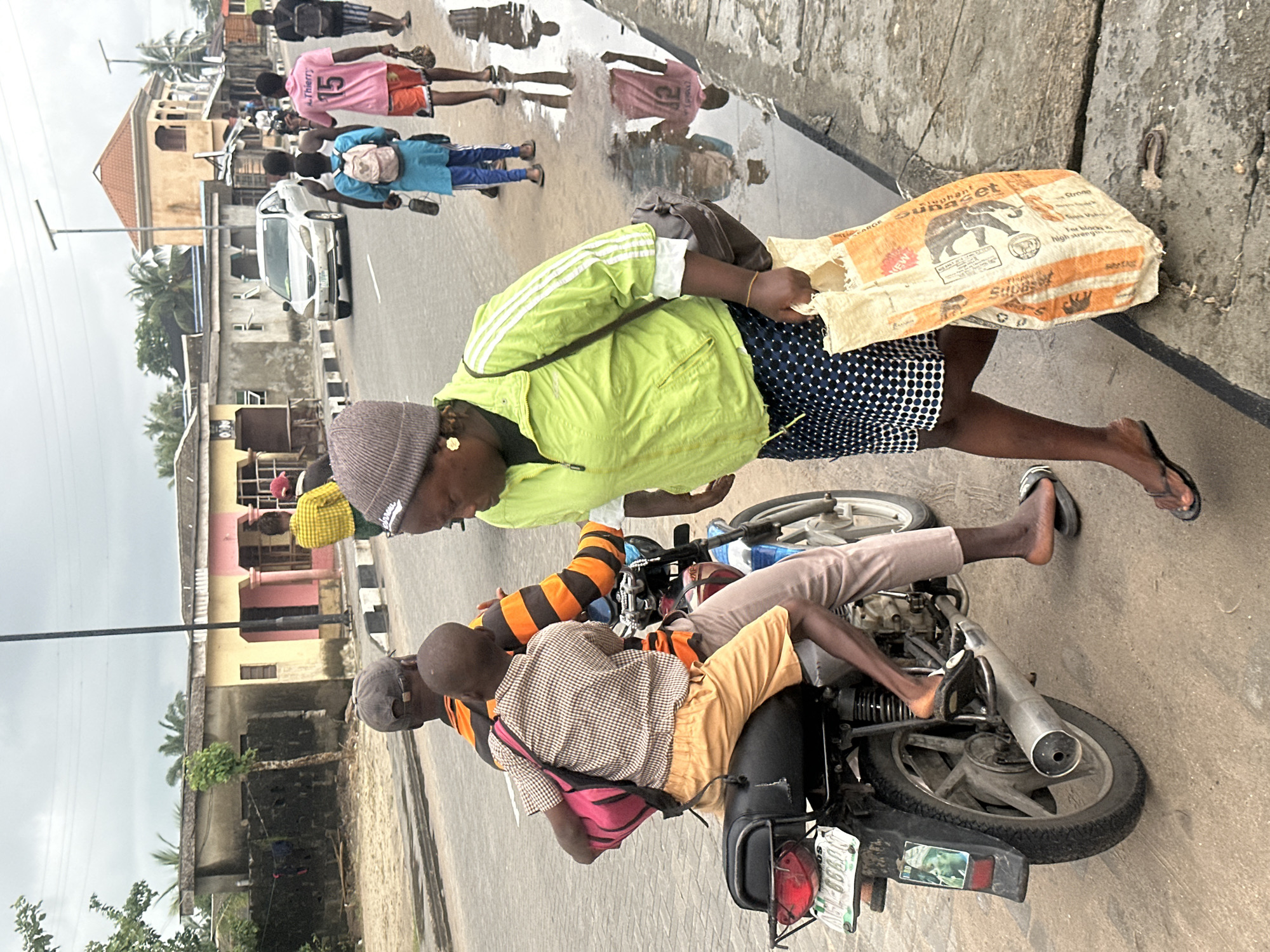 A street scene in Badagry, Nigeria, features a man and a boy on a motorcycle, with a woman walking beside them. The man, wearing a black cap and an orange and black striped shirt, is operating the motorcycle. The boy, positioned in front of him, is wearing a plaid shirt and tan shorts, with a pink backpack. The woman, in a bright green jacket and a blue and white polka dot skirt, carries a large, off-white woven bag with orange lettering. She is wearing a grey beanie. The background shows a street with buildings, palm trees, and a cloudy sky. Other people and a silver car are visible in the distance, suggesting a busy urban environment. The road appears wet, with reflections in puddles on the side.
