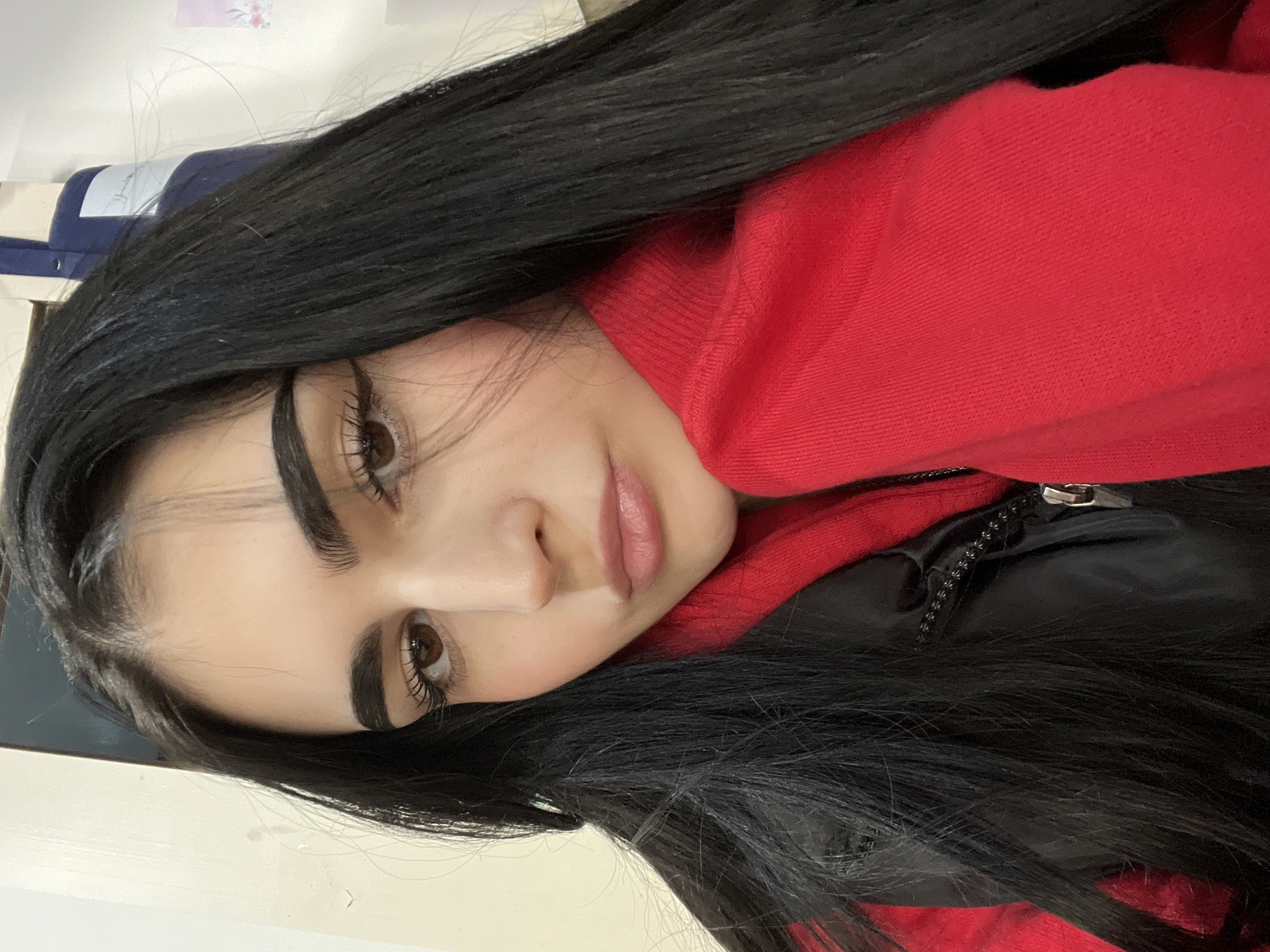 A close-up, rotated view shows a woman with long, straight, black hair. She is wearing a red hooded sweatshirt over what appears to be a black jacket. Her facial features include brown eyes, dark eyebrows, and light pink lipstick. The background is blurry but appears to include a white surface, a blue object, and a floral design. There is no information in the image to suggest the location is in Burgas, Bulgaria.