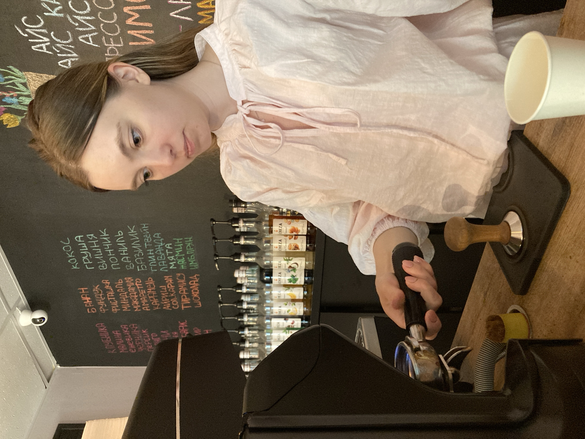 In Chaikovsky, Russia, a young woman with blonde hair, wearing a light pink ruffled blouse, is preparing a drink at a coffee shop. She is holding a portafilter in her right hand, engaged in the process of tamping coffee grounds. The background features a black chalkboard menu displaying various flavor options written in chalk, along with shelves lined with syrup bottles. To the right, a white paper cup sits on the wooden counter, and a tamper is placed on a black mat. A security camera is mounted on the ceiling.