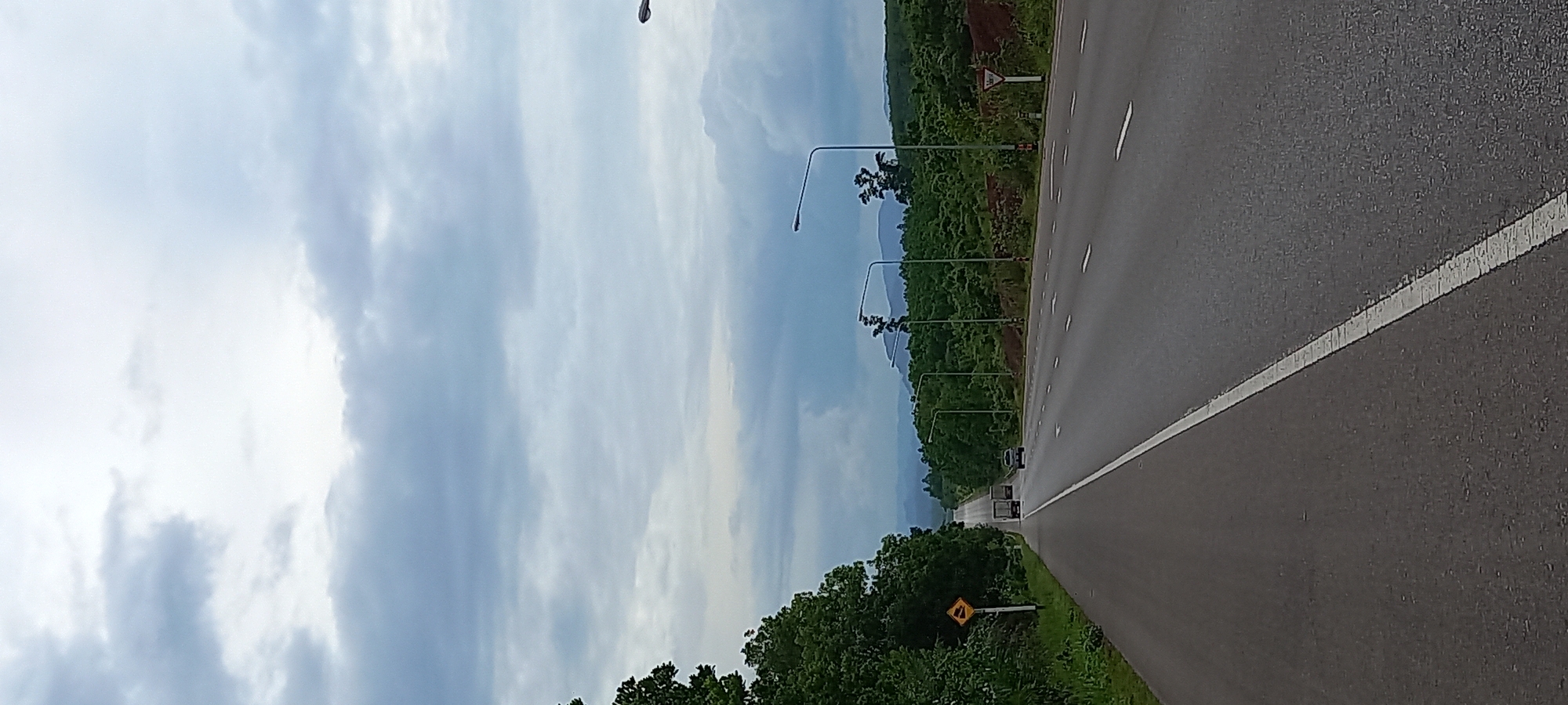 The image depicts a road in Khian Sa, Thailand. The road is asphalt with a white dividing line. The sky is cloudy. Trees and vegetation line the road. There are streetlights along the road. A yellow sign is visible on the side of the road, as well as another traffic warning sign farther down the road. In the distance, there are two cars traveling on the road.