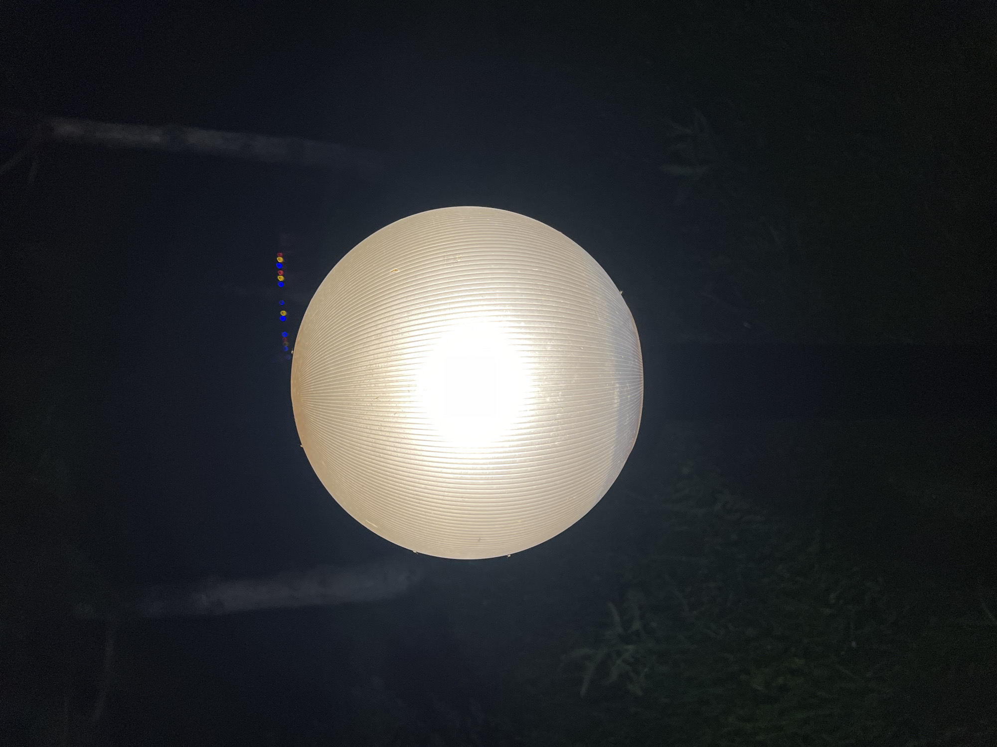 A spherical, translucent light fixture is illuminated from within, casting a bright glow against a dark background. The fixture has a ribbed texture. In the upper portion of the image, a faint trail of small, colorful lights can be seen. The lower portion of the image shows dark foliage and what appear to be two vertical, pale, indistinct shapes, possibly tree trunks or posts. The scene is dimly lit, suggesting it is nighttime or a very dark environment. The location of Plast Univska, Russia is noted. There are no visible people or specific actions taking place. The overall impression is of an outdoor light fixture at night.