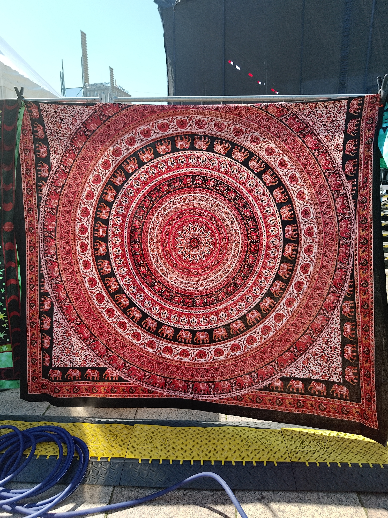 A tapestry with a concentric circular pattern primarily in red, black, and tan, with elephant motifs, is displayed on a metal frame outdoors, likely in an urban environment, though the specific location is Unknown, Unknown. The tapestry is hung against a backdrop of scaffolding, a black tarpaulin, and the sky. A coiled blue hose rests on a yellow and gray textured surface, possibly a cable protector, in the foreground. The scene is lit by natural sunlight.