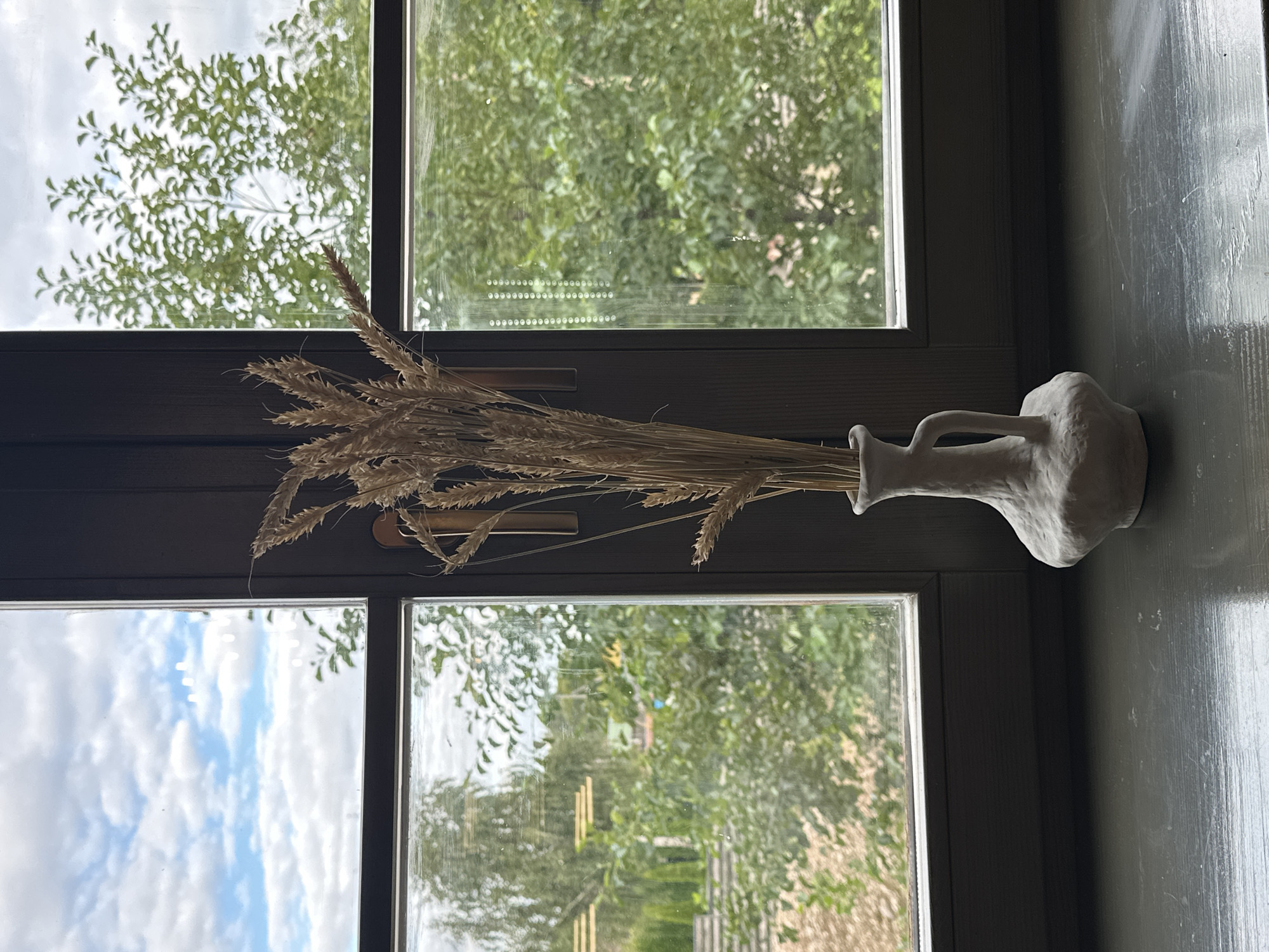 The image displays a still life composition on a dark gray windowsill, set against a backdrop of a window with a view of greenery and a cloudy sky. In the center, a white, irregularly shaped ceramic vase holds a bunch of dried wheat stalks. The vase has a small, curved handle. The window frame is dark, dividing the view into multiple panes. Through the glass, lush green trees and foliage are visible, suggesting an outdoor environment, possibly a garden or park. The sky above is blue with scattered white clouds. The windowsill itself appears to be made of wood and shows some signs of wear. The overall lighting suggests it is daytime, with natural light filtering in from the window.