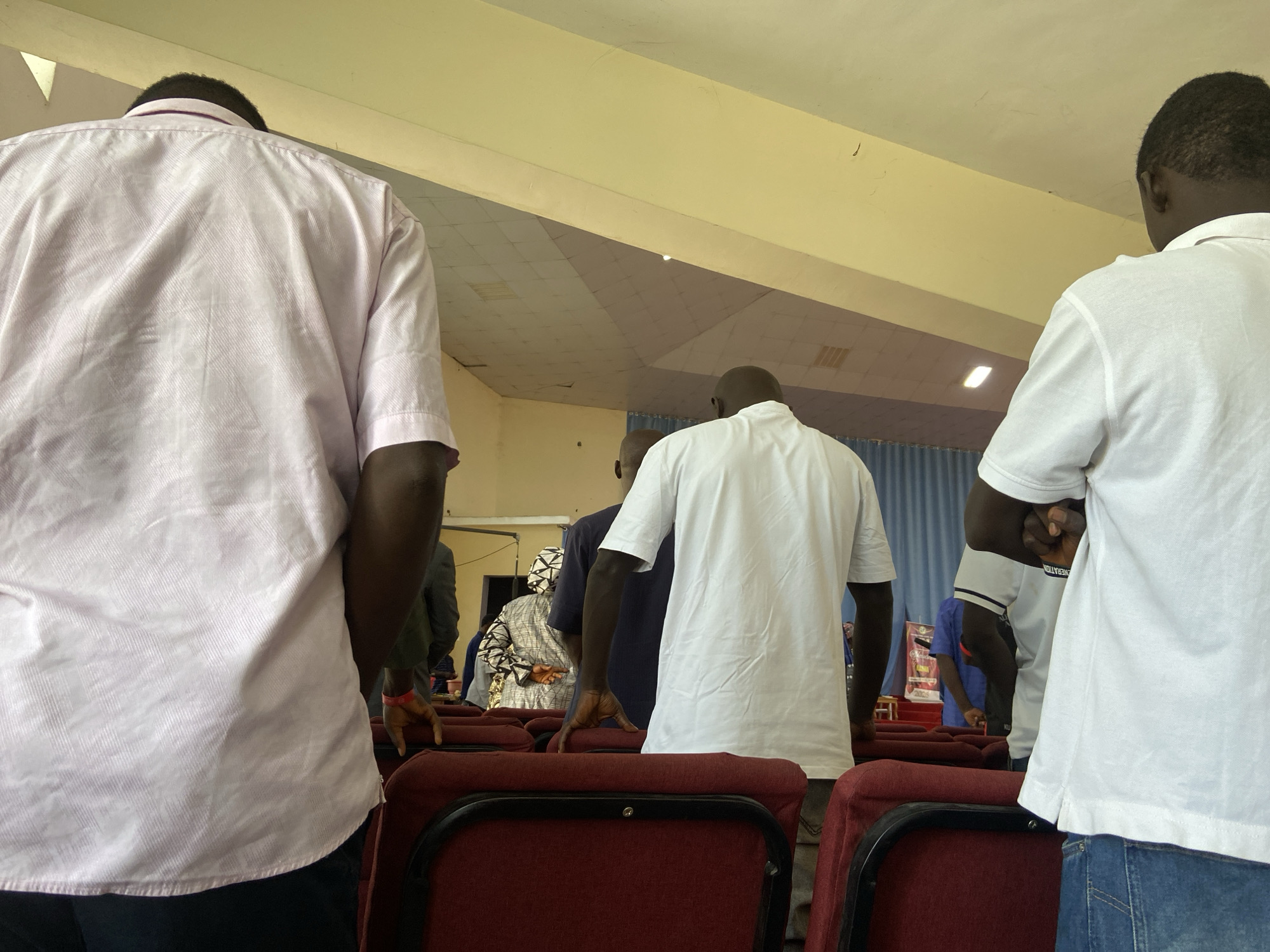 This image captures an indoor scene within the city of Mararaba, Nigeria, depicting a gathering from a rear perspective. The foreground features the backs of three men. On the left, a man wears a light pink, short-sleeved collared shirt and dark trousers. His hands are not visible. In the center, a bald man wears a white, long-sleeved tunic, with his right hand resting on a red upholstered chair back. On the right, another man wears a white, short-sleeved polo shirt and blue jeans, with his right arm bent at the elbow, hand out of frame.
Several rows of red upholstered chairs with black metal frames extend into the mid-ground. Between these chairs, other individuals are standing. To the left-center, a man in a dark blue short-sleeved shirt leans forward. Behind him and to the left, a person wearing a patterned grey and white garment and a head covering is partially visible, facing away. Further in the mid-ground, various individuals, some in lighter shirts or more formal attire, are seen standing or partially obscured.
The ceiling is light yellow with visible light-colored ceiling panels, and a prominent yellow structural beam traverses the upper portion of the frame. A rectangular light fixture is visible on the ceiling in the background. Blue curtains adorn the far back wall, behind which a raised platform or stage is partially visible. On this stage, a banner with red and white elements is present, with some text, possibly "GENERATION 2026," partially discernible. The overall lighting suggests a well-lit indoor environment. All individuals are standing, facing towards the front of the room, suggesting active participation in an event.