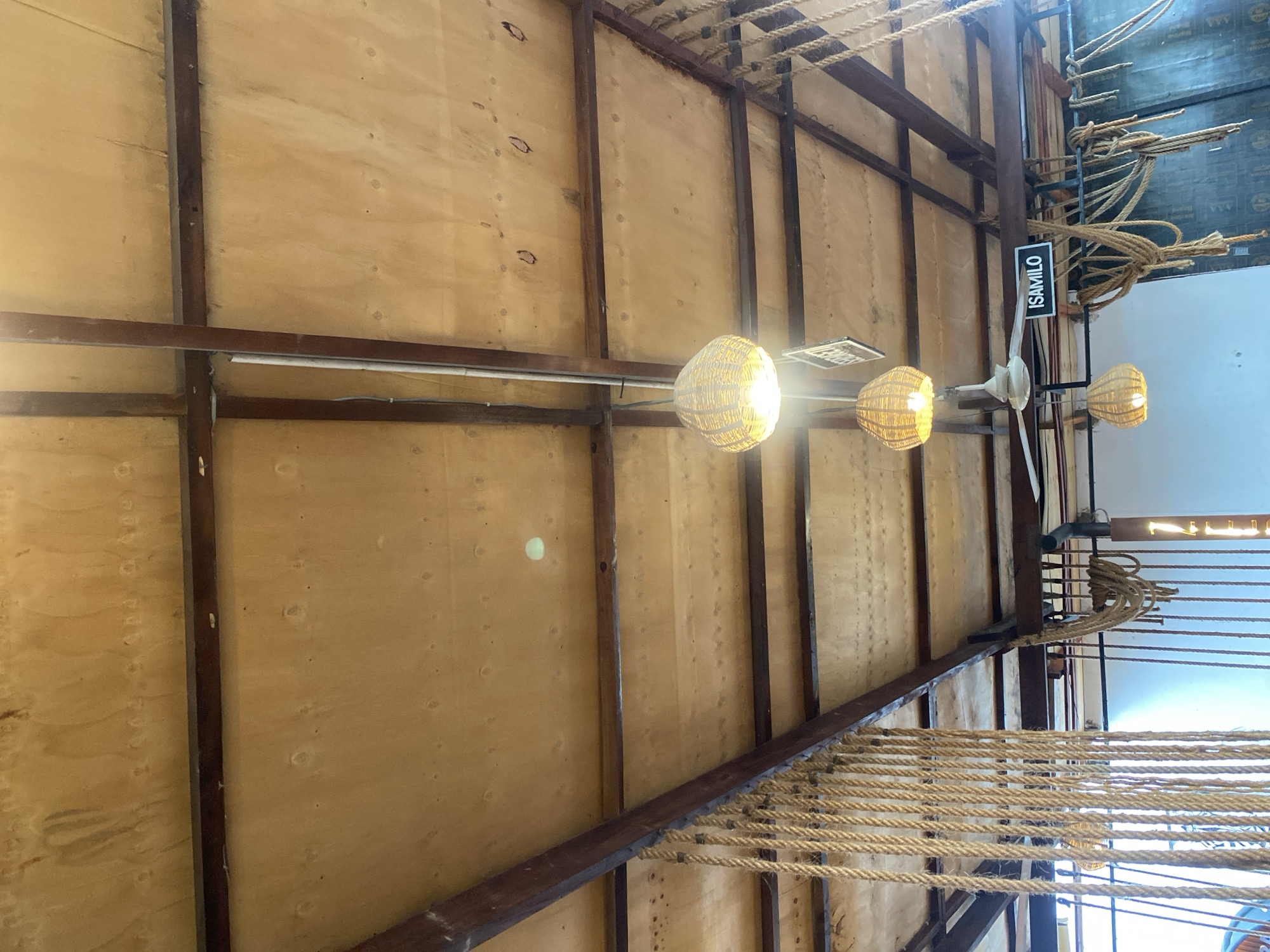 The image shows the interior of a building in Dar es-Salaam, Tanzania. The building has a high ceiling with exposed wooden beams and plywood panels. Several hanging lights with woven shades are visible. A rope structure is suspended from the ceiling. A sign that says "ISAMILO" is seen hanging by a fan.
