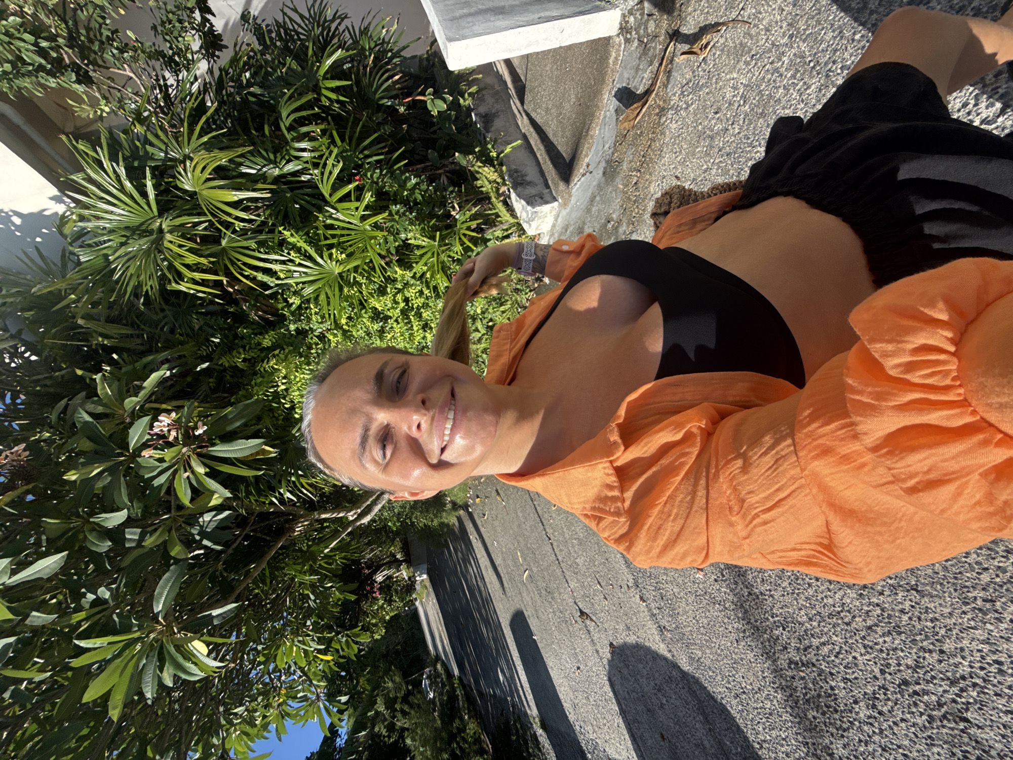 A woman is taking a selfie on a paved road, possibly in Ko Samui District, Thailand, given the lush tropical foliage and sunlight. She is smiling and holding her hair. She is wearing an orange open shirt over a black bra and black shorts. The road is sloped and casts shadows from trees and plants. Sunlight is illuminating the scene.
