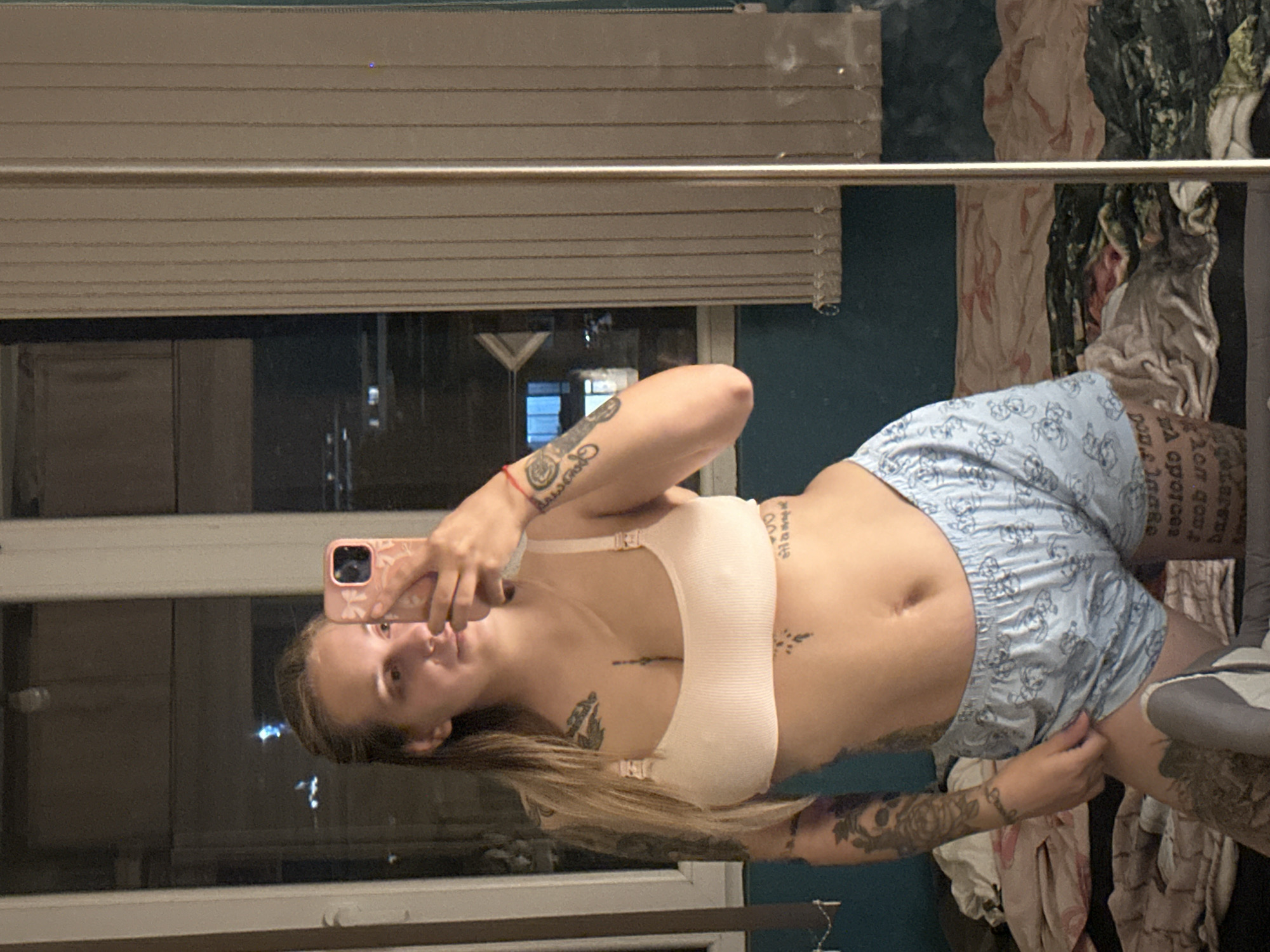 A young woman with blonde hair is taking a mirror selfie. She is wearing a light pink bra and blue pajama shorts with a cartoon print. She has tattoos on her arms, torso, and legs. She is standing in what appears to be a bedroom, with a bed in the background. The room has a window and vertical blinds. It is nighttime, and lights from the city are visible through the window. The location is not specified.