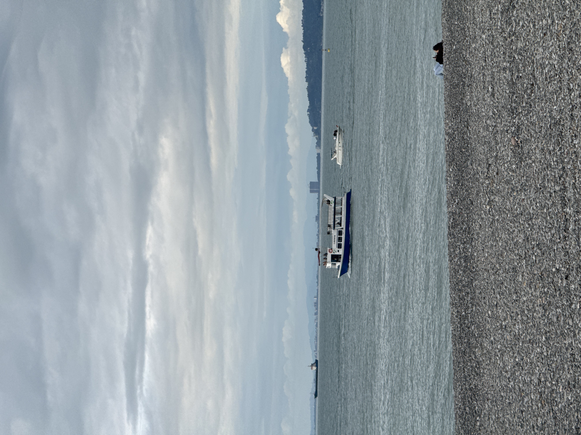 The image shows a view of the Black Sea from the pebble beach in Batumi, Georgia. The sky is overcast with a variety of cloud formations. Two boats are visible on the water. One boat is larger and painted with blue and white colors, with some people on board. A smaller white boat is further away in the background. Across the water, a thin strip of land with trees and buildings can be seen. A person is sitting on the pebble beach in the upper right corner. A large pebble beach is visible.