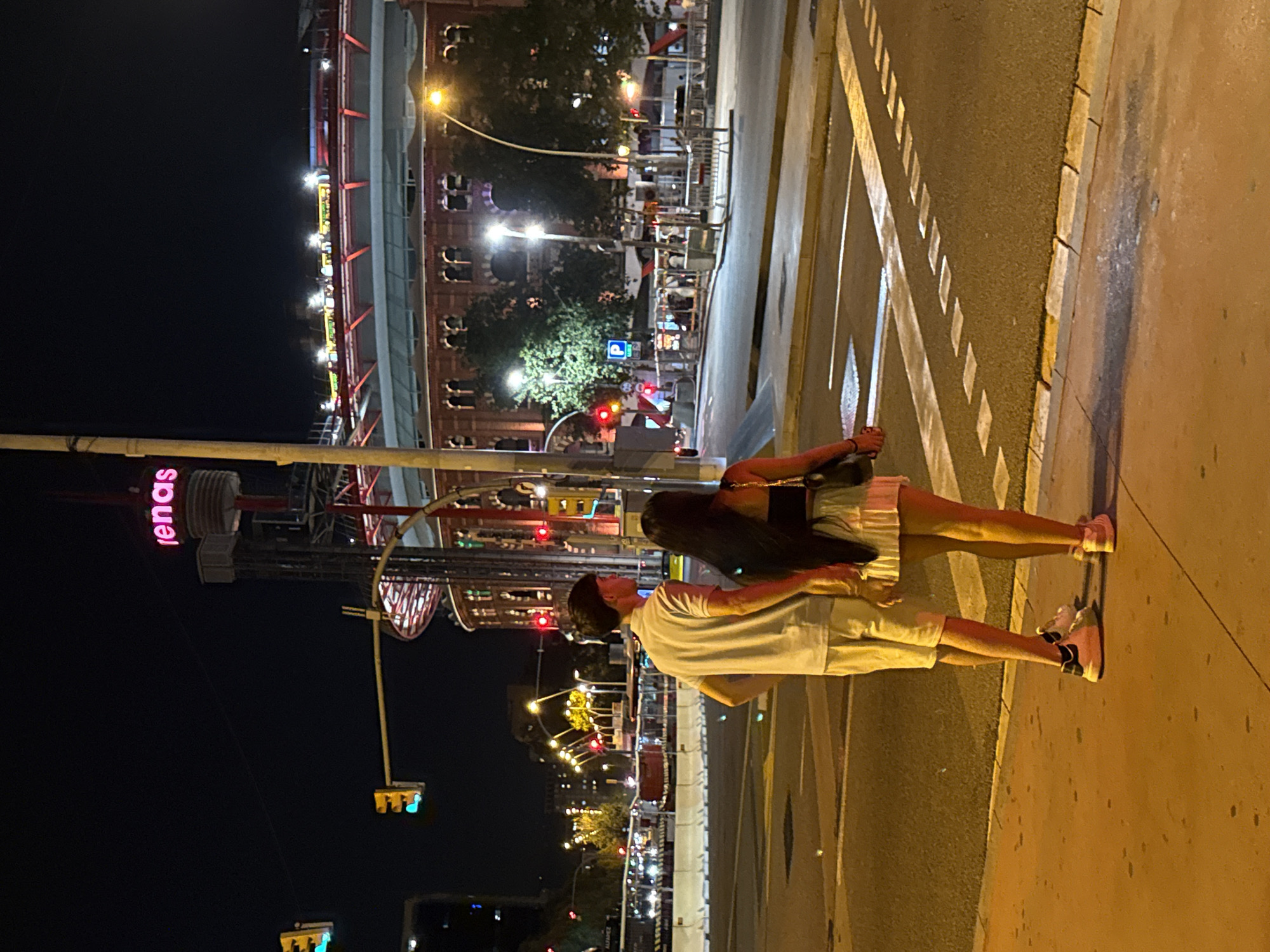 A man and a woman are standing on a street corner in Barcelona, Spain, at night, holding hands. They are facing away from the camera, looking towards a large, circular, brick building with arched windows, illuminated by lights. Above the building, the word "arenas" is visible in neon pink letters. A traffic light is red, indicating they should wait to cross. The street is paved, with a crosswalk marked by white lines. The surrounding area is dimly lit, with streetlights and lights from buildings and vehicles visible in the background.