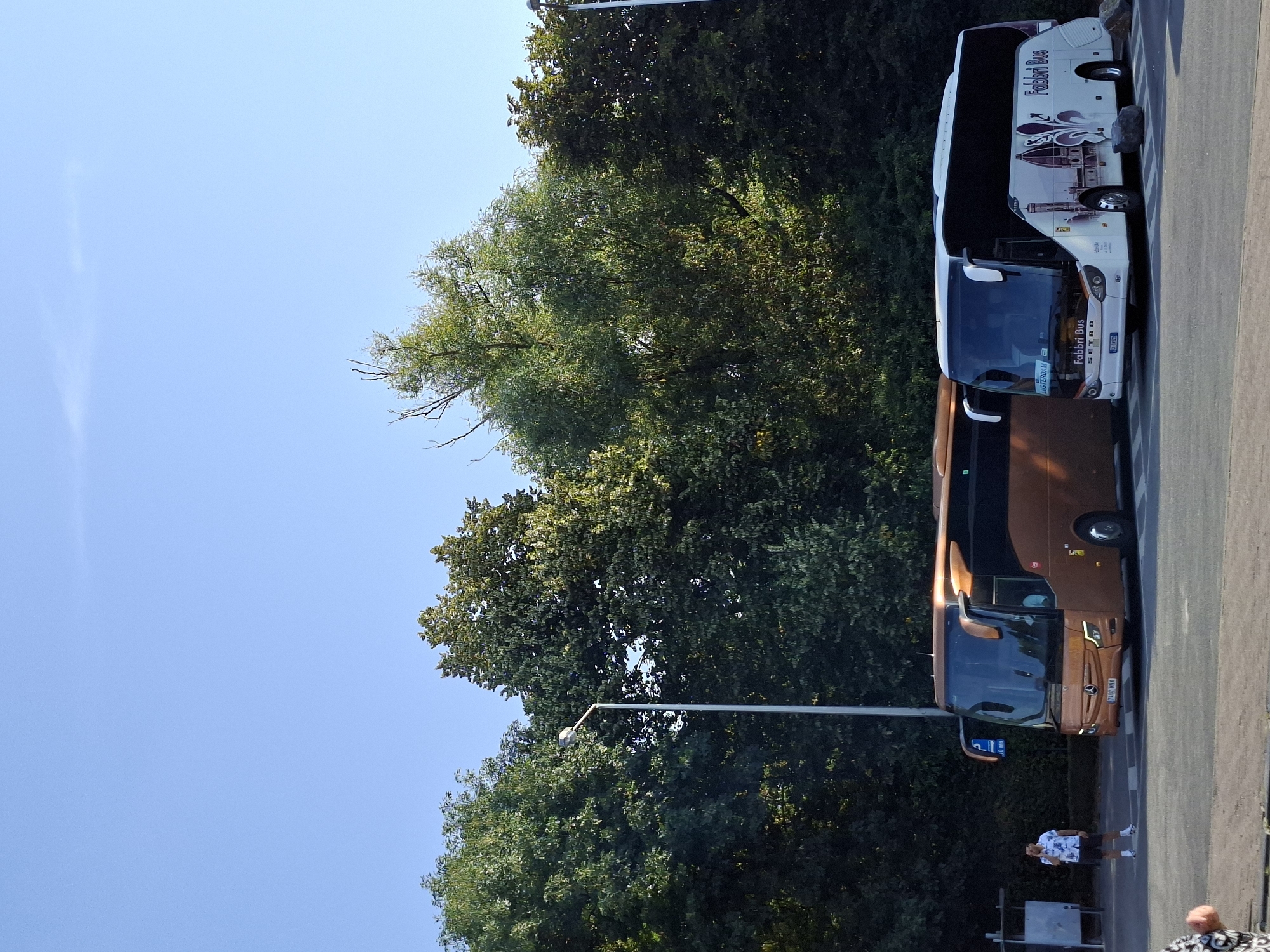 Two buses are parked in a lot with trees in the background. The bus on the left is brown and the bus on the right is white with a graphic of the Duomo of Florence and the Fabbri Bus logo. A man in a floral shirt and shorts is standing near the brown bus, talking on his phone. The scene appears to be an outdoor parking area, possibly at a bus station or a tourist attraction, given the presence of two tour buses. The sky is clear blue. The license plate on the brown bus reads "7467 MNI". The license plate on the white bus reads "CE 669TD". A sign on the white bus reads "AMSTERDAM".