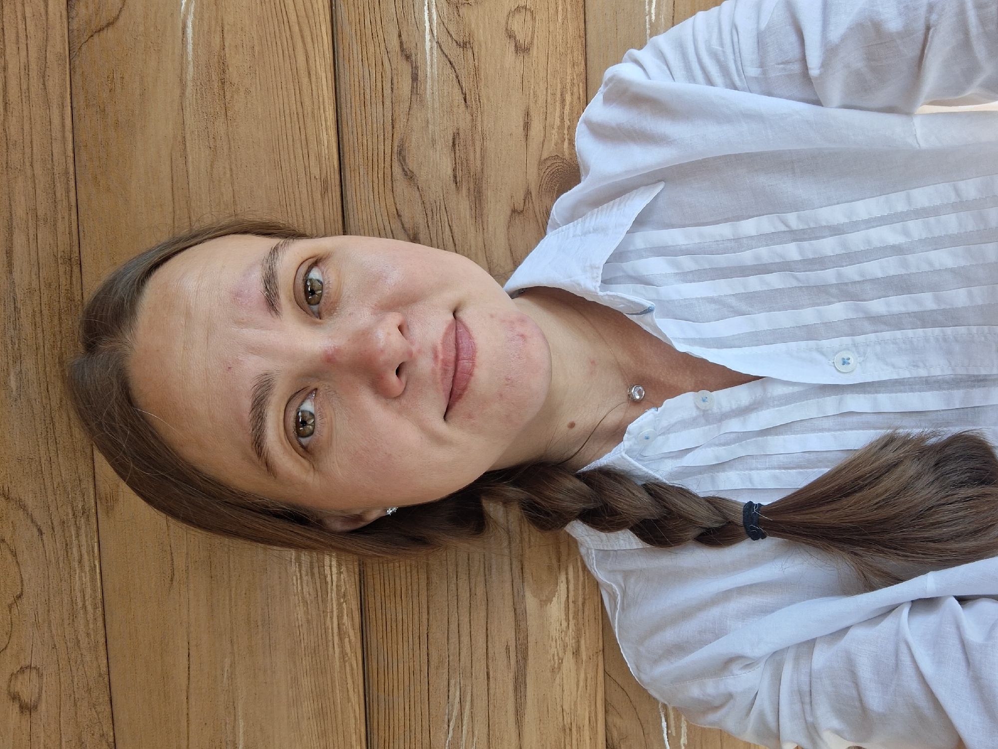 A young woman with brown hair braided into a single plait, wearing a white button-down shirt with subtle pleating detail, is lying on a wooden surface. Her hair falls across her shoulder and chest, secured with a dark hair tie. The wooden planks appear to be outdoors, suggesting a natural setting. Her expression is relaxed, with a slight smile. A small, shimmering earring is visible in her ear. The lighting suggests it could be daytime, with natural light illuminating the scene. The overall impression is one of casual comfort.