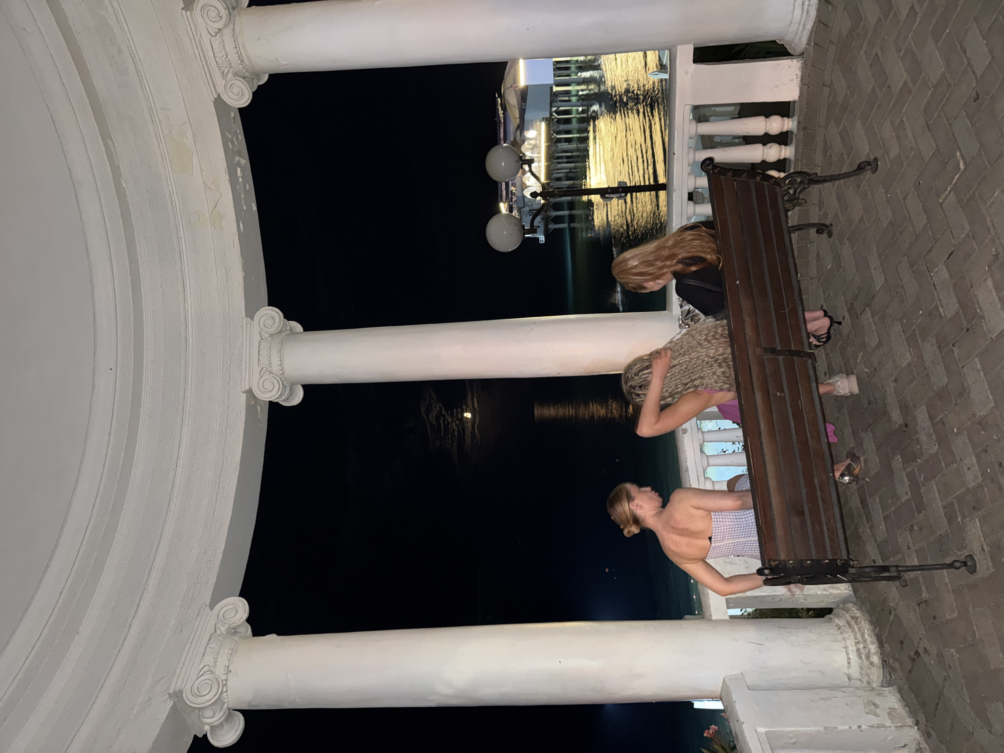 Three young women are seated on a wooden bench under a white rotunda at night in Sochi, Russia. The woman on the left is looking to her left, wearing a strapless gingham dress. The middle woman is facing away, with her arm raised and touching her head, her curly blonde hair is prominent. The woman on the right is also facing away, with long blonde hair. Behind them, a full moon is visible over a calm body of water, reflecting the lights of a building with a pier. The rotunda has large white columns and a curved, white ceiling. The ground is paved with brick.