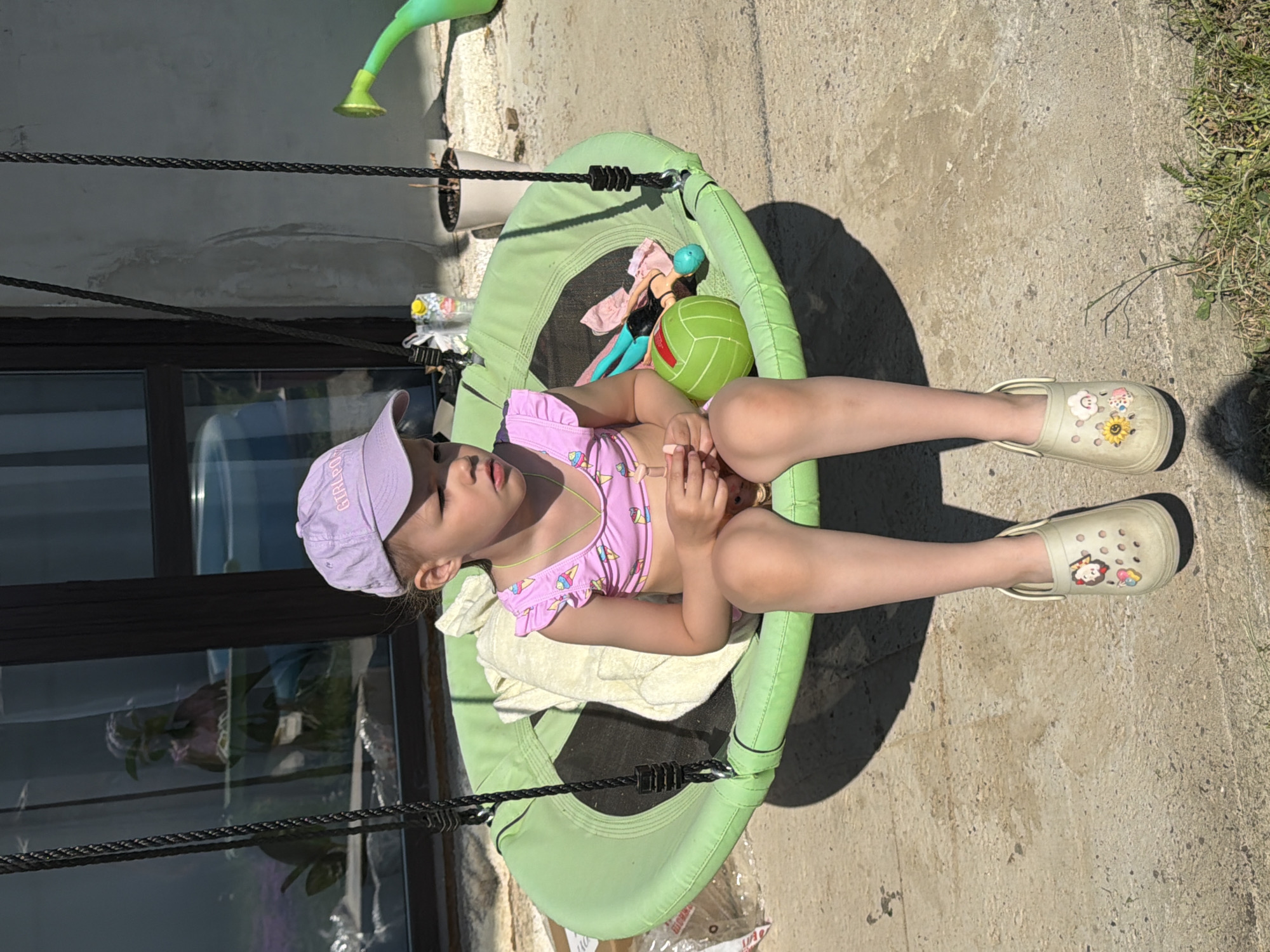 A young child is lying on a green, swing-like trampoline. The child is wearing a light purple cap, a pink and purple swimsuit with ice cream cone designs, and light-colored shoes. The child is lying on a white towel. Beside her on the trampoline is a green ball and a doll. The location is outdoors, possibly in a yard, with a concrete surface and some grass. A building with windows is visible in the background. The city of Chaikovsky, Russia, is mentioned.