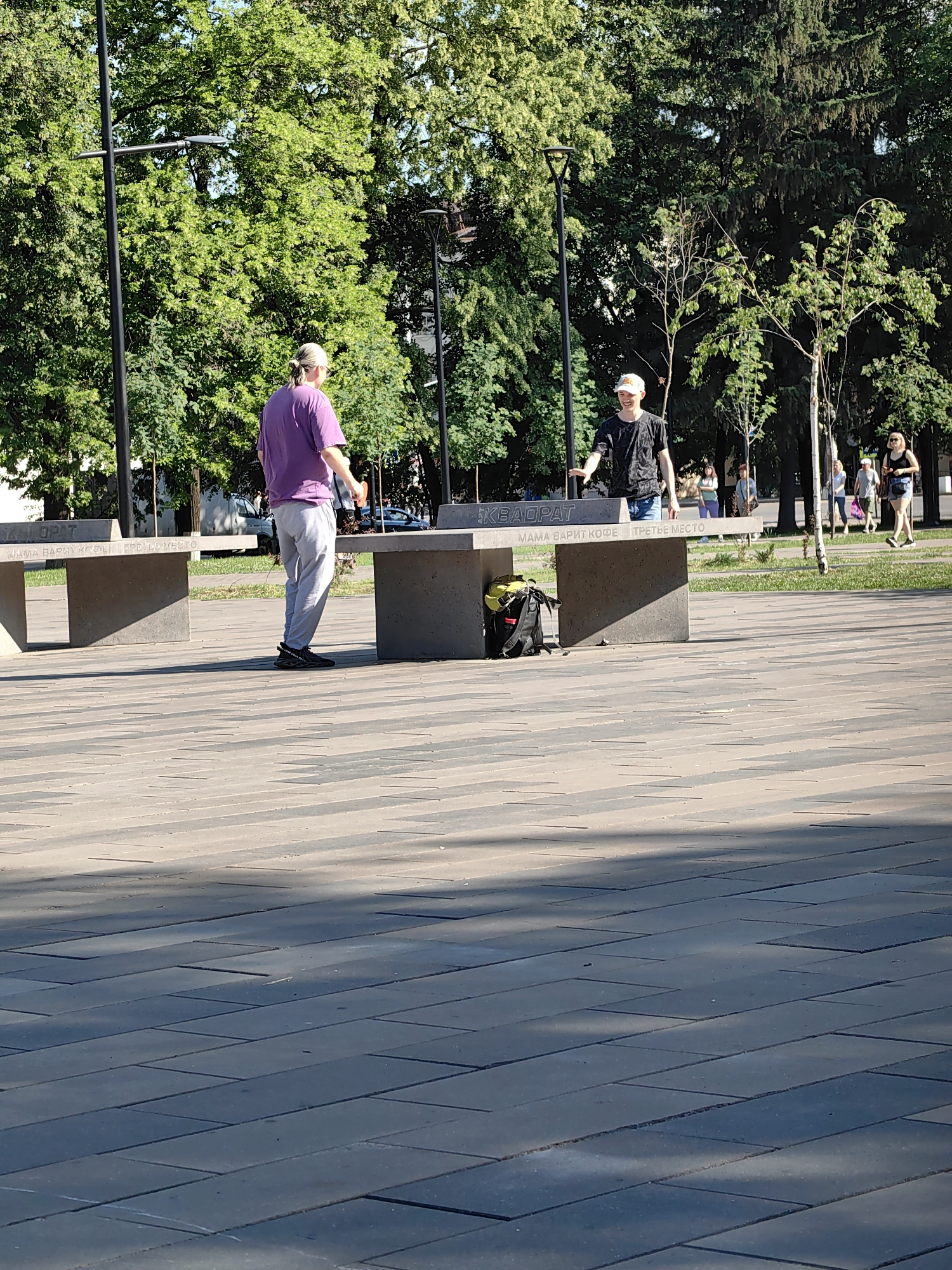 The media file shows an outdoor scene in a park on a sunny day. Two young men are standing on either side of a concrete table, possibly a ping pong table, and appear to be interacting. One man on the left is wearing a purple t-shirt and grey sweatpants, with his back partially turned to the camera. The man on the right is wearing a black t-shirt and blue jeans, with a white baseball cap. He is looking at the other man and has his hands raised as if in a playful gesture.
In the background, there are several trees with lush green foliage, suggesting it is a warm season. Other people are visible in the distance, walking and enjoying the park. The ground is paved with patterned tiles. There is a backpack with a yellow and black strap placed on the concrete table on the left.
Visible text on the concrete table reads "КВАДРАТ" and "МАМА ВАРИТ КОФЕ" on the left side, and "ТРЕТЬЕ МЕСТО" on the right side. The overall atmosphere appears casual and relaxed, suggesting a typical day in a public park. The sunlight casting shadows indicates it is daytime, likely mid-morning to afternoon. The location context is Ufa, Russia.