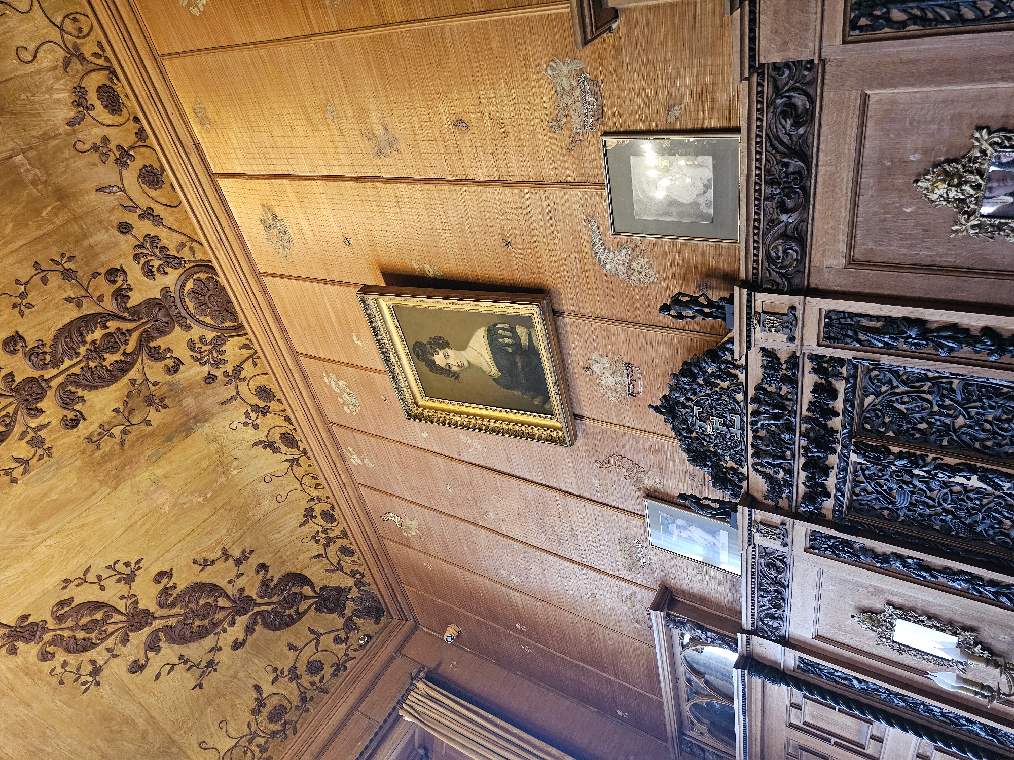 The image shows a corner of an ornately decorated interior, likely a room within a historic building. The location is potentially in Алупкинский городской совет, Ukraine. The corner is defined by walls and ceiling panels covered in wood.
The wall on the left is adorned with dark brown floral patterns on a light brown surface. A portrait in a gold frame hangs on the adjacent wall, which is constructed from narrow wooden planks. These planks also feature faint, lighter-colored floral patterns. Another framed picture is above and to the right of the portrait.
A complexly carved wooden structure is on the right, showing darker wood and intricate designs. Additional framed pictures are visible on this structure. The overall impression is of an elaborate, historical interior.