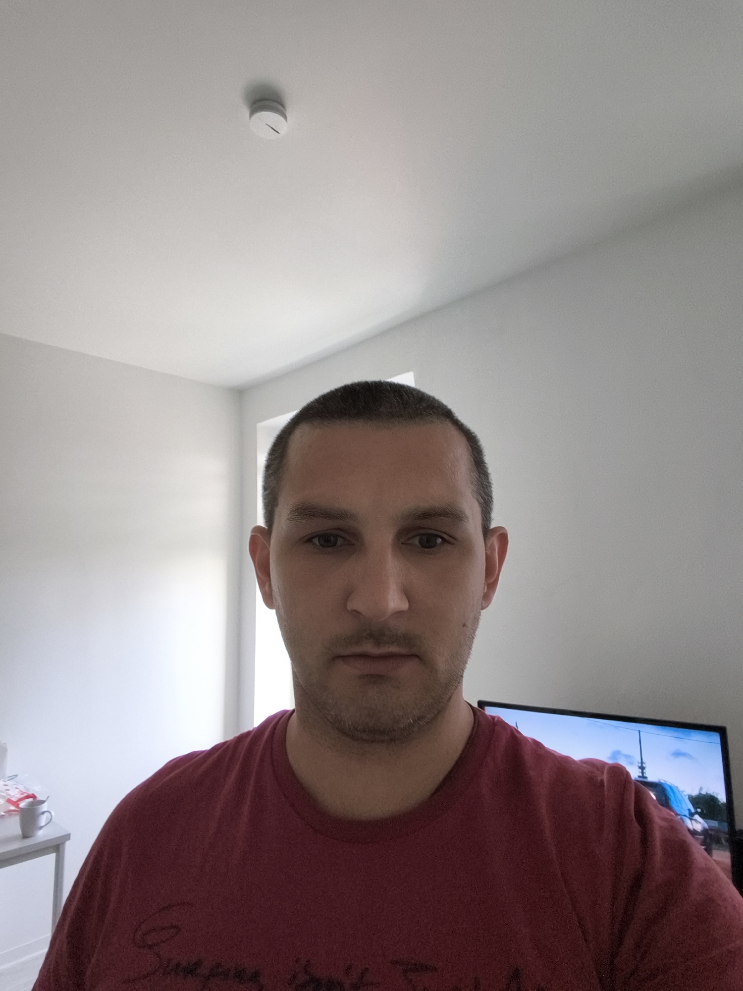 The image shows a head-and-shoulders shot of a light-skinned man with short dark hair and stubble. He is wearing a maroon t-shirt. The man is positioned in the center of the frame, looking directly at the camera. The background consists of a white wall and ceiling, with a white smoke detector visible on the ceiling. To the right of the man, a portion of a television screen is visible, displaying an image of a vehicle. To the left of the man, a gray table with a white mug on it is partially visible. The scene appears to be indoors, possibly in a residential setting. The image does not contain any specific landmarks or clues that would help determine its location within the city of Wolgast, Germany.