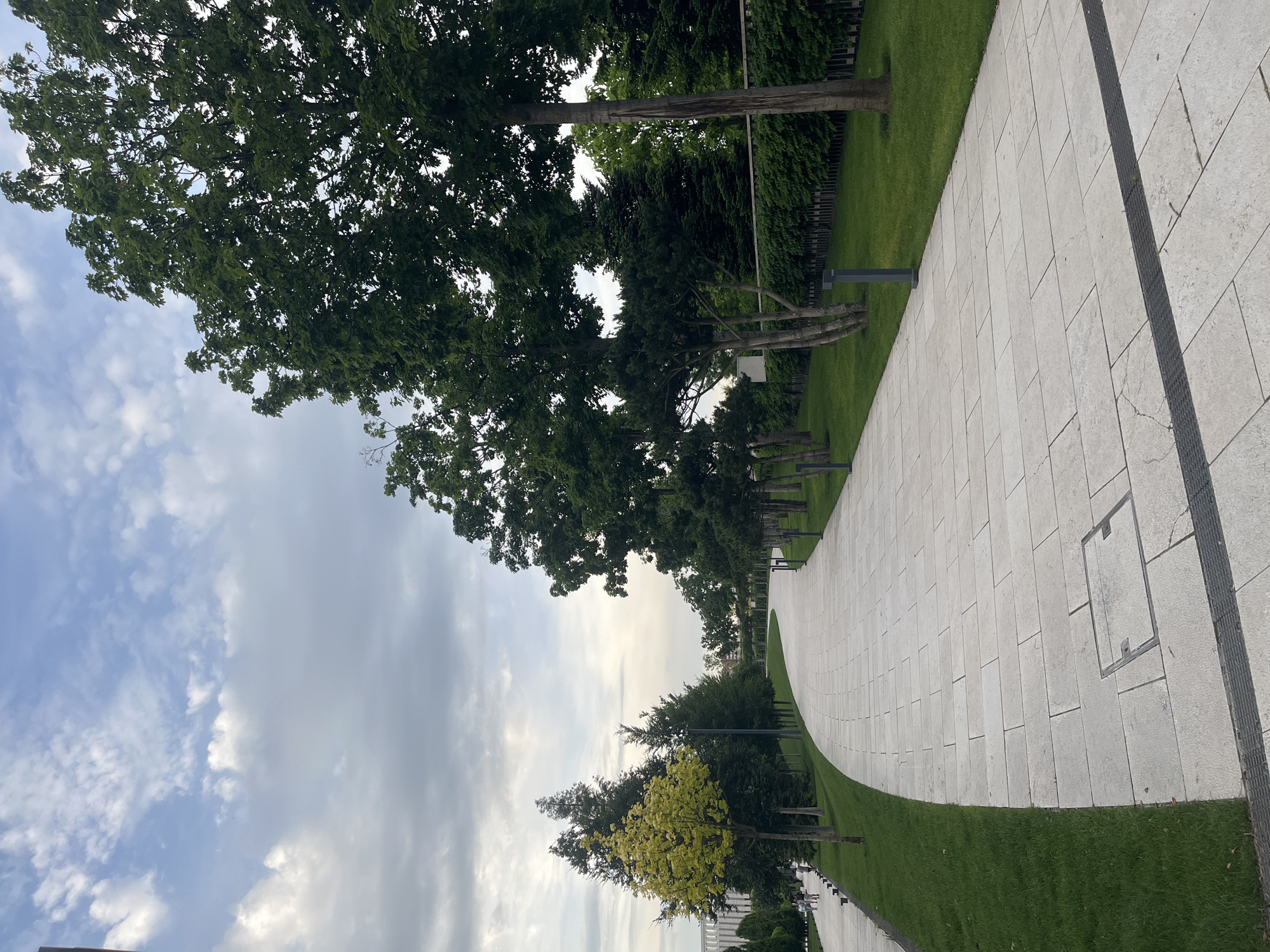 The image captures a serene park scene in Krasnodar, Russia, featuring a wide, light-colored paved pathway that curves gently from the foreground into the distance. The pathway is constructed from rectangular tiles and has a dark, textured strip running alongside it on the right, potentially for drainage or as a tactile surface. A square manhole cover with a metal frame is visible embedded in the path on the right.
Lush green grass borders both sides of the path, and numerous trees with dense green foliage line the edges of the park. One prominent tree in the mid-ground stands out with noticeably lighter, yellowish-green leaves. Modern, dark-colored bollard-style streetlights are intermittently placed along the path's edge on the right.
The sky overhead is partly cloudy, displaying a mix of bright white and softer grey clouds, indicating a daytime setting, possibly late afternoon. In the far distance, faint figures of pedestrians can be discerned on the path, suggesting the park is a public area frequented by people. The overall scene conveys a sense of calm and well-maintained urban green space. There is no visible text in the image.