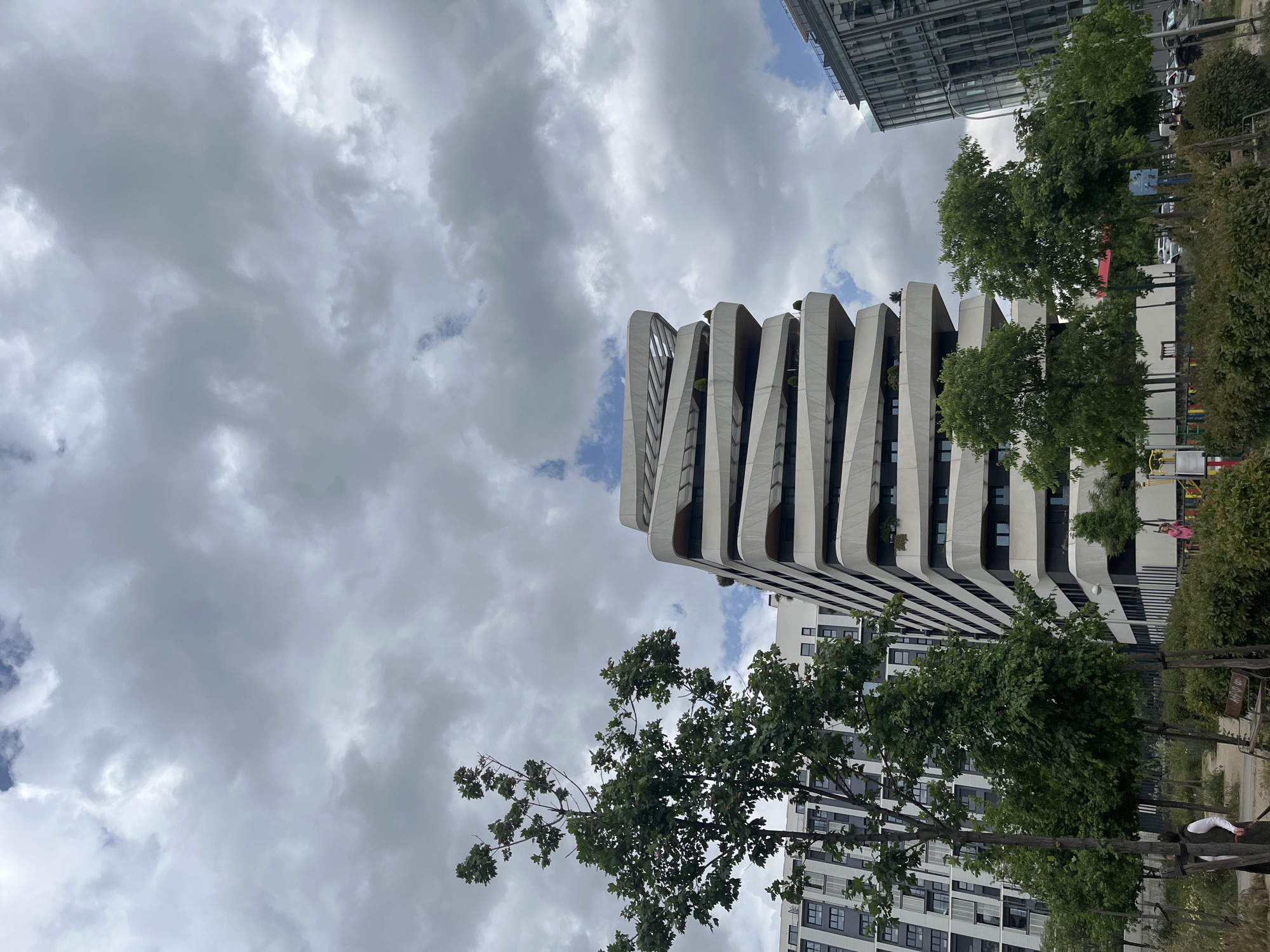 The image presents an urban landscape dominated by modern architecture and natural elements under a cloudy sky. A distinctive, tall building with a unique wavy, layered facade, featuring integrated balconies or terraces with small plants, stands prominently. To its right and top, a sleek, glass-fronted skyscraper is partially visible.
In the foreground and mid-ground, numerous trees with dense green foliage are dispersed, alongside bushes and other landscaping. A paved area or street, with several parked cars, runs along the right side of the frame. Adjacent to the trees on the right, there's a children's playground with a red slide and yellow climbing equipment. A person wearing a white top is partially visible near the playground area at the bottom right. Another, less distinct building with dark-framed windows is situated in the bottom left.
The sky is largely overcast with large grey clouds, although several patches of blue sky are visible, indicating it is daytime. The image appears to have been captured in portrait orientation but is displayed rotated counter-clockwise. Text on distant signs is unreadable, except for a faint "HAM..." on one element. The overall scene suggests a contemporary urban or suburban setting, aligning with the specified location of Madrid, Spain.