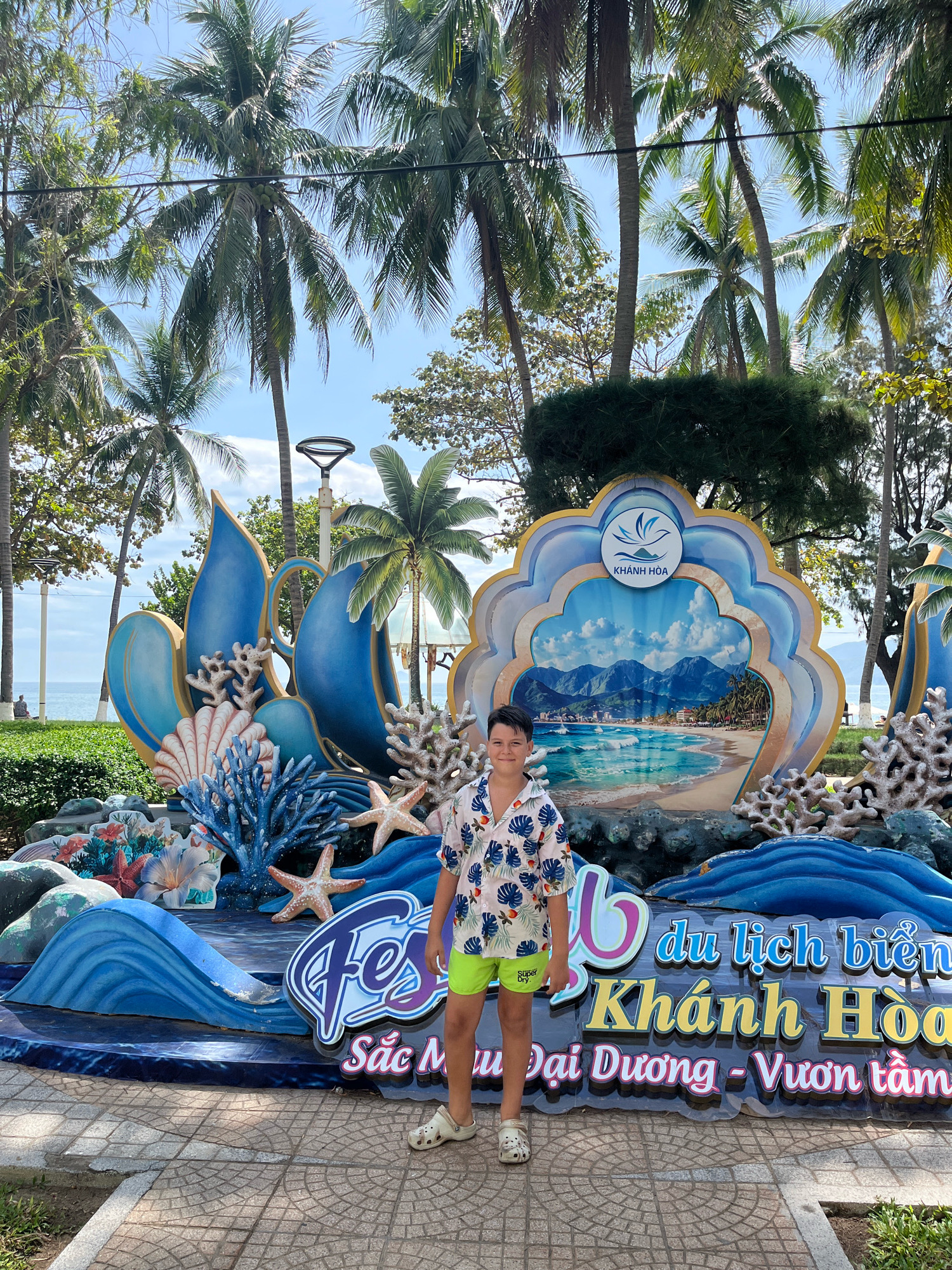A young boy stands in front of a large decorative display featuring a beach scene with mountains in the background. The display has a scalloped edge and is adorned with blue wave-like elements, coral, and seashells. Text in Vietnamese is visible on the display, indicating it is related to "du lịch biển Khánh Hòa" (beach tourism of Khánh Hòa). The boy is wearing a short-sleeved floral shirt and green shorts. Palm trees are visible in the background, suggesting a tropical or coastal location. The scene appears to be outdoors, possibly in a public area like a park or promenade in Nha Trang, Vietnam, given the text referencing Khánh Hòa province, which includes Nha Trang. The sky is blue with some clouds.
