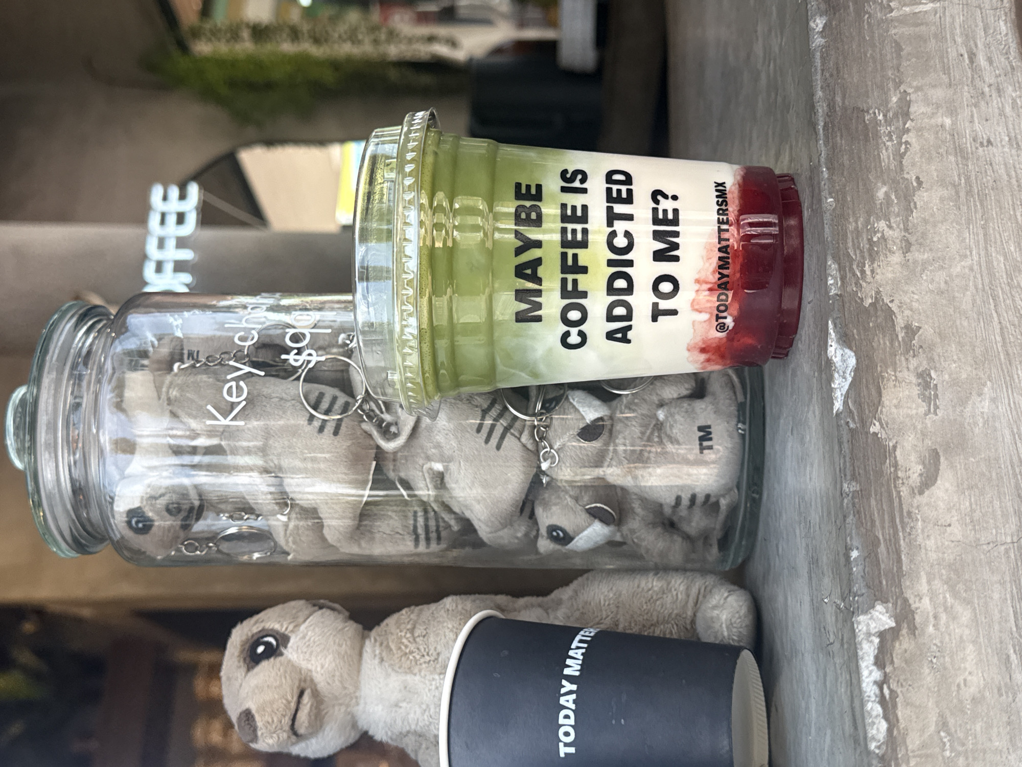 A still life shot captures a vibrant, layered beverage alongside various merchandise on a concrete surface, likely outside a coffee shop. The image is rotated clockwise by 90 degrees.
In the foreground, a tall, clear plastic cup holds a three-layered drink: a light green layer on top, a milky white layer in the middle, and a red (possibly strawberry) layer at the bottom. The cup features black text that reads, "MAYBE COFFEE IS ADDICTED TO ME?" and "@TODAYMATTERSMX".
Next to the drink, a clear glass jar is filled with numerous small, light brown plush keychains, resembling meerkats or raccoons, complete with silver rings. Partially visible text on the jar includes "Key-ch" and "$90", possibly indicating "keychain" and a price. A "TM" symbol is also visible on one of the plush toys inside the jar.
Below the jar and beverage, a larger, soft plush toy, similar to the keychains, lies on the grey concrete. Adjacent to it is a black paper cup with white text that reads, "TODAY MATTER".
In the blurry background, a sign with "COFFEE" (partially visible as "IFFEE") suggests the setting is a coffee shop. The bright lighting indicates it is daytime. The concrete surface appears worn and textured.