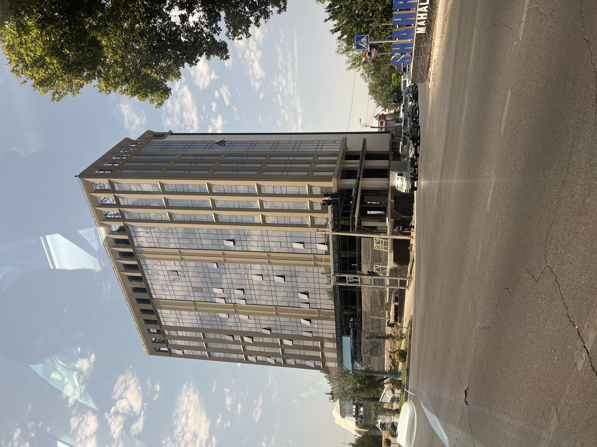 The image shows a modern, glass-fronted office building in Tashkent, Uzbekistan. The building, under construction on lower floors, features a cream-colored exterior frame and numerous windows, some of which are open. In the foreground, a cracked asphalt road runs in front of the building, with several cars parked or driving. To the right, a sign with the words "SHAHRI" and "MAHAL" is visible, along with a pedestrian crossing sign. Trees line the street and the sky is partly cloudy. Reflections of the sky and what appear to be car interiors are visible on the building's glass facade, suggesting the photo was taken from inside a vehicle.