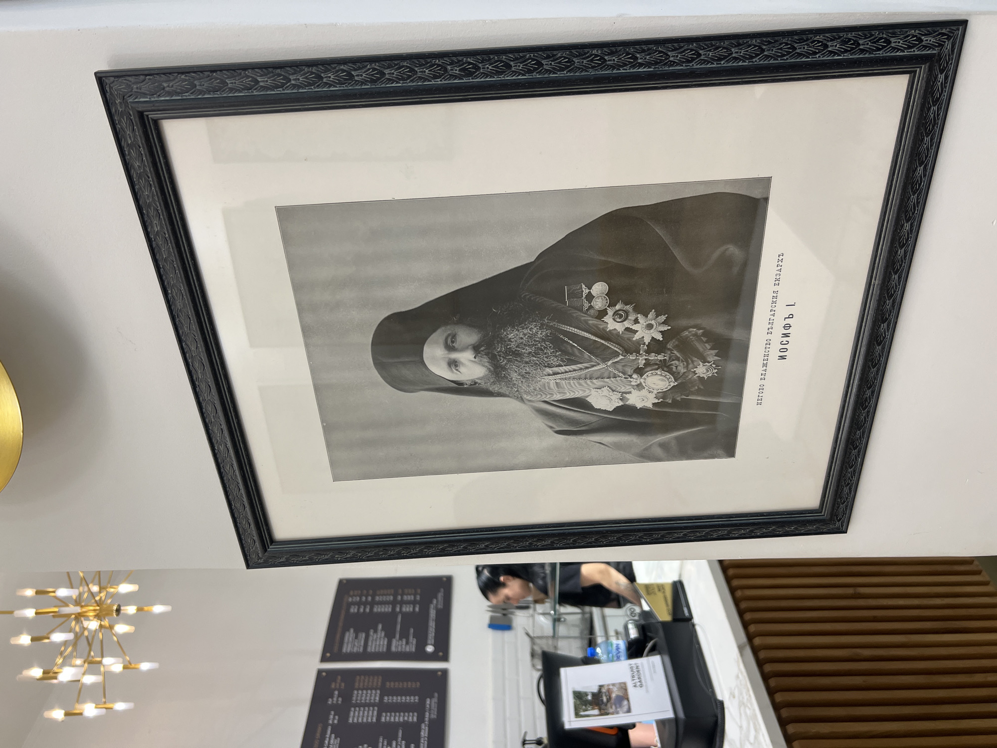 A black and white framed portrait of a man with a long beard, wearing a dark clerical hat and robe adorned with medals, hangs on a white wall. Below the portrait, text is visible in what appears to be Cyrillic script. To the left of the framed portrait, a modern chandelier with multiple lights hangs from the ceiling. Further to the left, a counter area of a cafe or restaurant is visible, with menus displayed on the wall and a woman standing behind the counter. The overall environment appears to be an interior space with a contemporary aesthetic, likely a commercial establishment. Based on the Cyrillic text and context of portraits of historical figures, this image was likely taken in Sofia, Bulgaria.