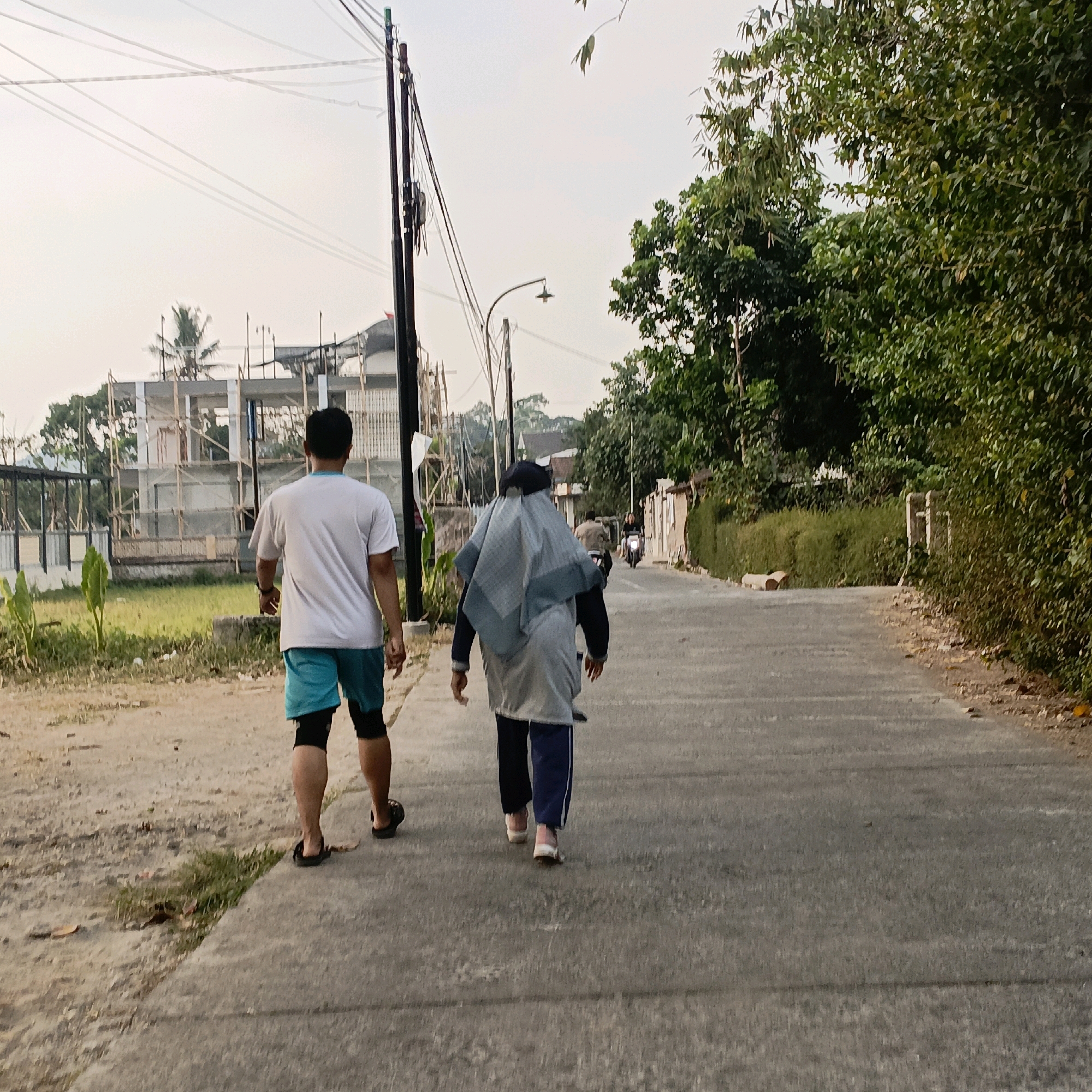 Two individuals are walking on an asphalt road in what appears to be a semi-rural area of Kesongo, Indonesia. The person in the foreground to the left is a male wearing a white t-shirt, turquoise shorts, and black knee-high socks with black sandals. The person to the right is wearing a light blue hijab, a dark long-sleeved top, and dark pants with light blue stripes, and light pink shoes. They are walking away from the camera, towards a curved section of the road.
In the background to the left, a multi-story building under construction is visible, with scaffolding and exposed framework. A palm tree stands near the construction site, and a green grassy area with some plants is present. To the right of the road, dense green trees and foliage line the path. Overhead, power lines are strung between poles, and a street lamp is visible. The sky is overcast and light. Further down the road, another person on a motorcycle is approaching. The overall scene suggests a normal day with people engaged in walking, and construction activity in the vicinity.