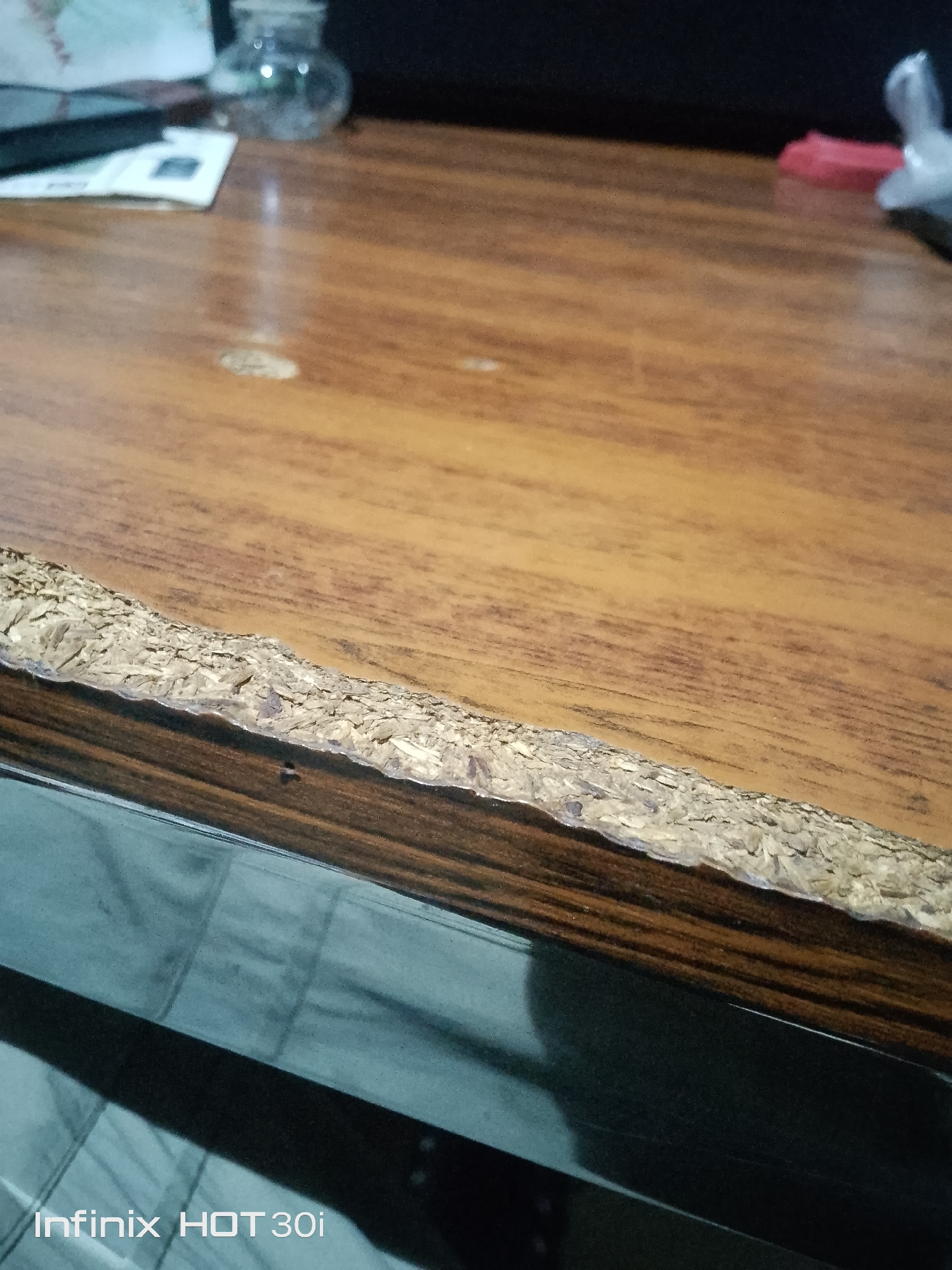The image shows a close-up view of a damaged particleboard edge on a wooden surface. The particleboard is peeling and chipped, revealing the fibrous material underneath. The wooden surface has a wood grain pattern and appears to be a tabletop or desk. In the background, out of focus, are some objects, including a clear glass jar and a portion of a black electronic device. The bottom of the image shows a reflective surface, possibly glass or a polished tabletop, with a shadow cast upon it. The text "Infinix HOT 30i" is visible in the lower left corner of the image, indicating it may be a screenshot or a photo taken with that device. The scene is indoors, with artificial lighting.