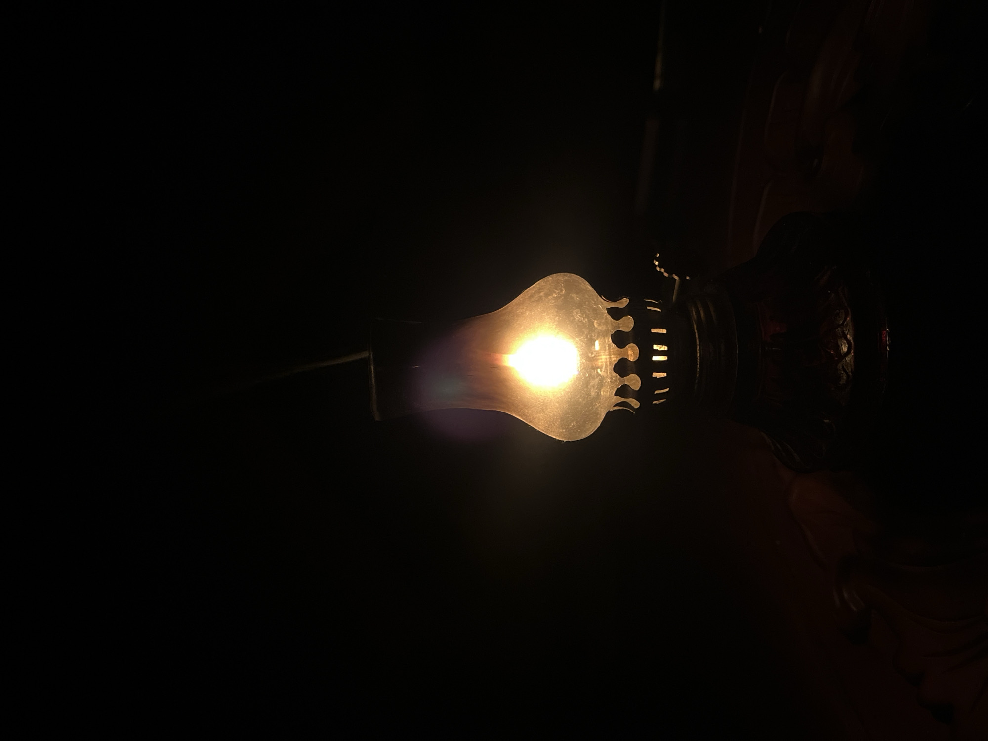A kerosene lamp illuminates a dark space. The lamp is oriented horizontally, with the bulbous glass portion to the left, and the metal fixture with decorative elements to the right. The scene is very dimly lit. The location is unknown.
