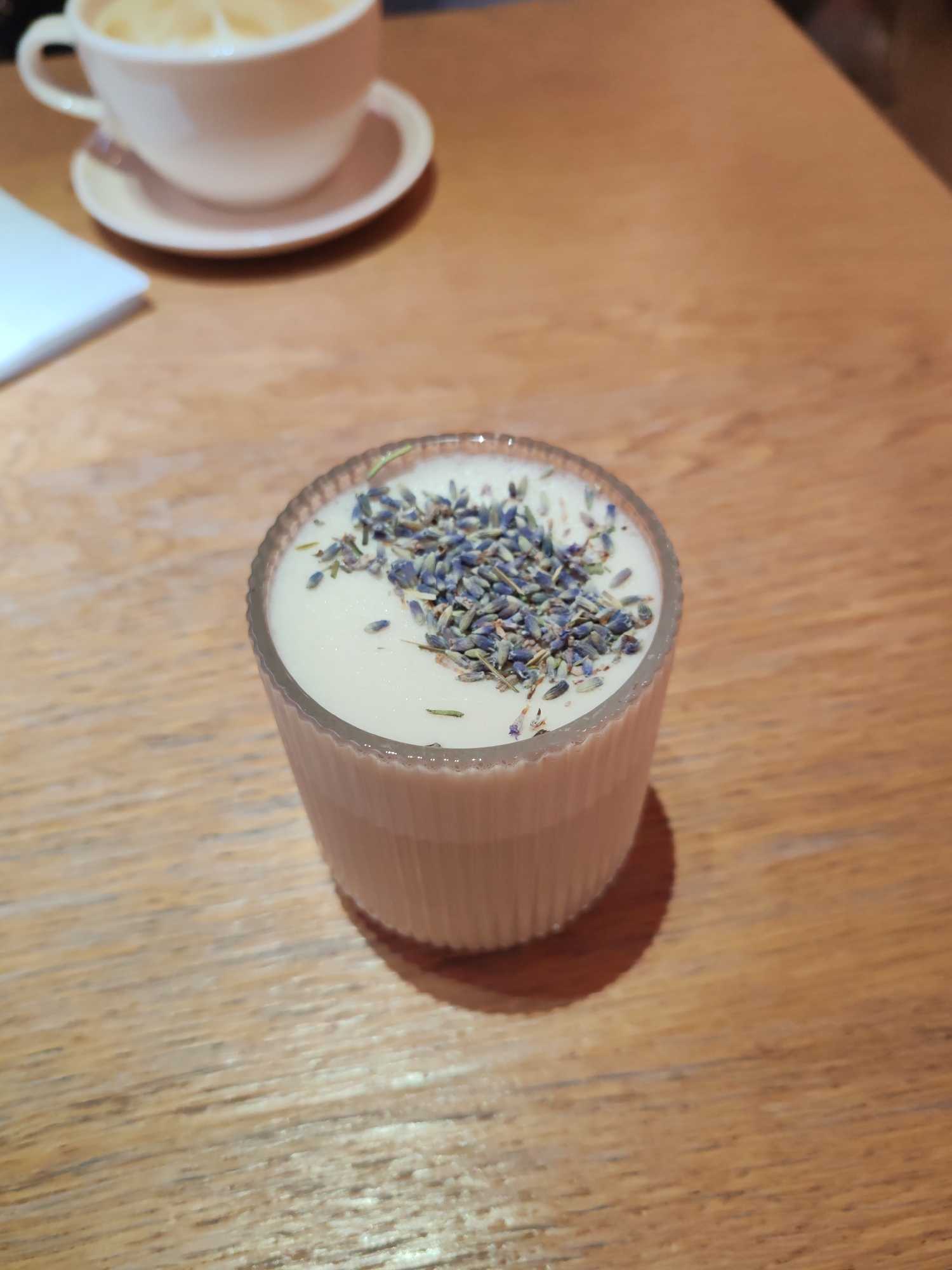 The image displays a close-up, high-angle shot of a light-colored drink or dessert in a clear, ribbed glass, placed on a wooden table. The glass holds a creamy, white substance topped generously with dried lavender florets, which are a mix of blue, purple, and grey hues, along with some small green specks. In the blurred background, to the upper left, a white ceramic cup with a handle rests on a matching saucer, appearing to contain a light-toned beverage, possibly a latte. A folded white napkin or paper is also partially visible on the left side of the background. The surface beneath the items is a light-toned wooden table with a distinct grain pattern. The lighting is soft and diffused, characteristic of an indoor setting such as a cafe or restaurant in Saint Petersburg, Russia. No individuals are visible, and there is no discernible text within the frame. The scene conveys a calm atmosphere, focusing on a pleasant refreshment.