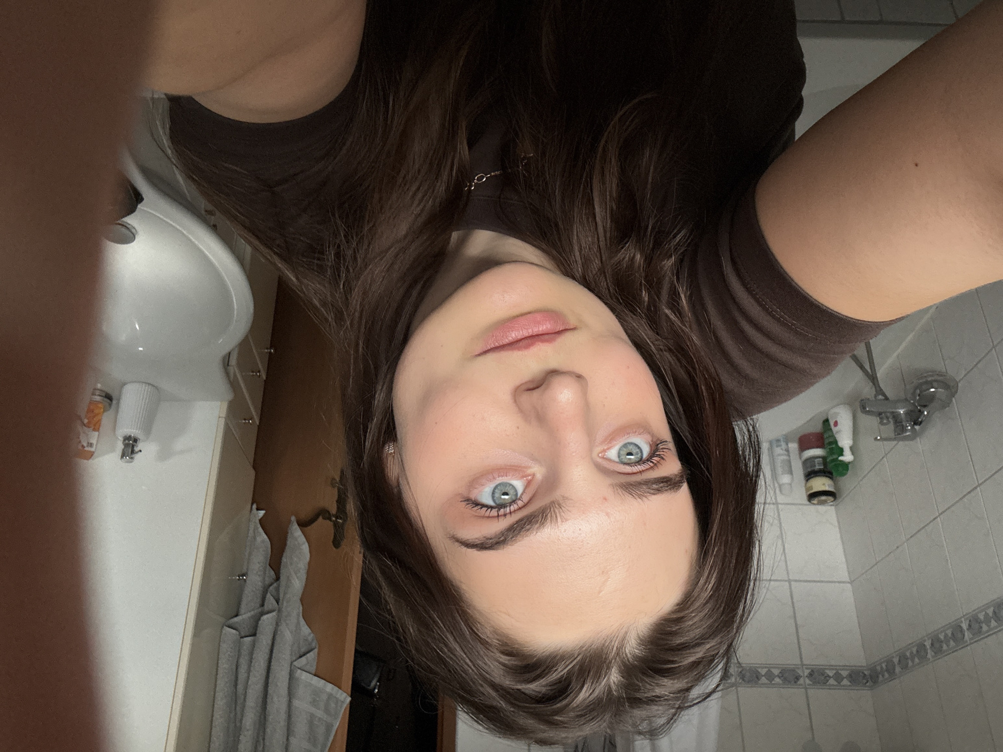 A close-up, high-angle selfie shows a young woman in what appears to be a bathroom. The image is significantly rotated counter-clockwise, presenting the woman's face upside down relative to the background elements. She has long, dark brown hair that frames her face, striking light blue eyes looking directly at the viewer, and a neutral, calm expression. Her lips are lightly colored with a pink hue. She wears a simple dark brown top.
The background reveals typical bathroom features. On the left, a white ceramic sink with a faucet is visible. To the right and behind her, white tiled walls with a decorative grey-patterned border can be seen, along with a metal faucet and various toiletries (including a white ceramic soap dispenser) on a shelf or ledge. Below the shelf, grey towels hang on hooks, next to what appears to be a wooden doorframe. The setting is indoors, under artificial lighting, with no indication of the time of day or weather. There is no visible text in the image. The location context provided is Hainburg an der Donau, Austria.