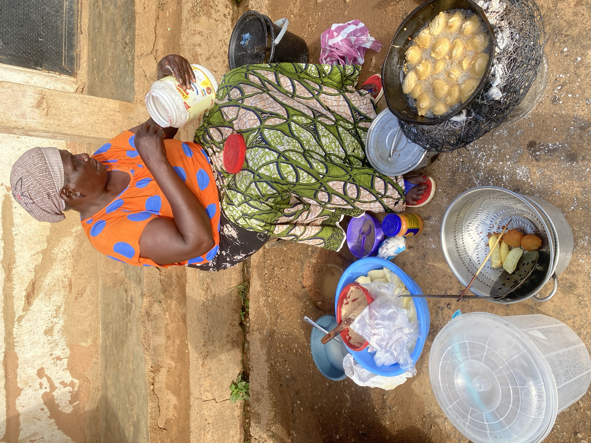 An overhead medium shot captures a woman, likely in Jos, Nigeria, seated outdoors and preparing food. She wears an orange t-shirt with blue polka dots and a patterned wrap skirt. Her head is covered with a beige and brown headscarf. In her right hand, she holds a large white plastic tub with a yellow label, and appears to be adding something from it. In front of her are various cooking implements and ingredients. A large blue bowl contains what appears to be dough and possibly some cut fruit or vegetables, wrapped in plastic. A red bowl with a wooden spoon sits inside the blue bowl. Next to it are smaller purple and blue containers. To the right, a black bucket with a pink and white cloth is visible. A large pan filled with oil and golden-brown fritters is placed over a charcoal brazier, with some of the fritters being fried. A metal colander holds some cooked fritters and skewers. A large clear plastic tub with a lid is situated to the left. The ground is composed of dirt and sand. The background features a rough, textured wall and a dark metal door. The scene suggests a food preparation area for sale or consumption.