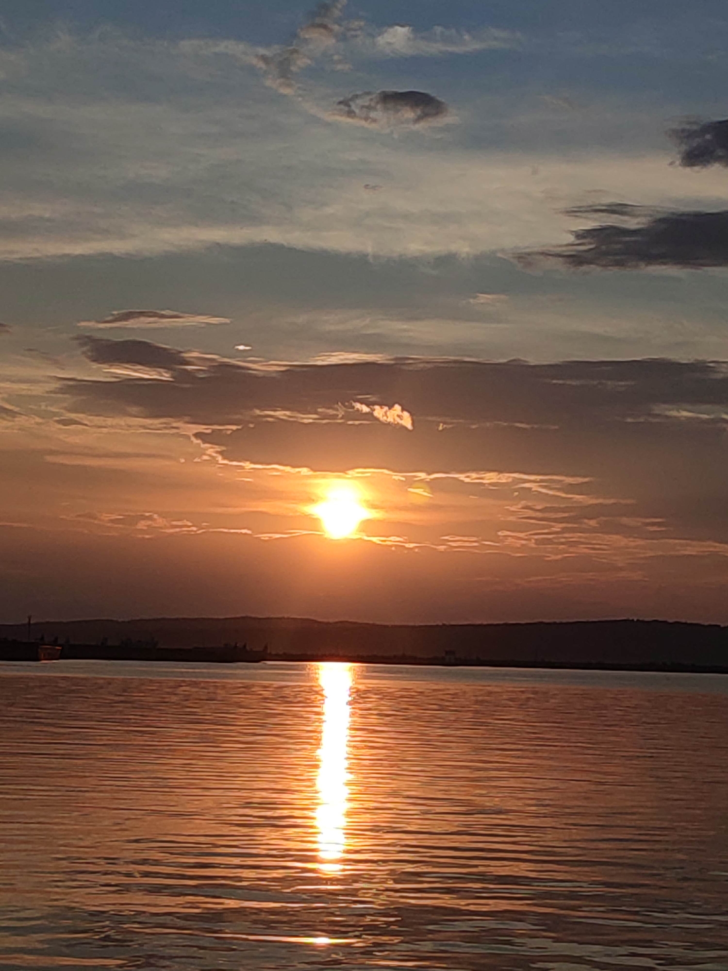 This is a vertical, eye-level, full shot of a sunset over a body of water. The sun is a bright, glowing orb in the center of the frame, slightly to the left, peeking through scattered, wispy clouds. The sky transitions from a pale blue at the top to a warm orange and yellow near the horizon, reflecting the sun's light. Thin, horizontal clouds are visible across the sky, some dark and silhouetted, others tinged with orange. The horizon line is a dark, indistinct silhouette of a distant landmass, possibly a shoreline with trees or buildings. The water in the foreground is calm, with gentle ripples creating streaks of reflected sunlight, predominantly in hues of orange and gold, with some darker, shadowed areas. The overall impression is one of serene natural beauty as day transitions into night.