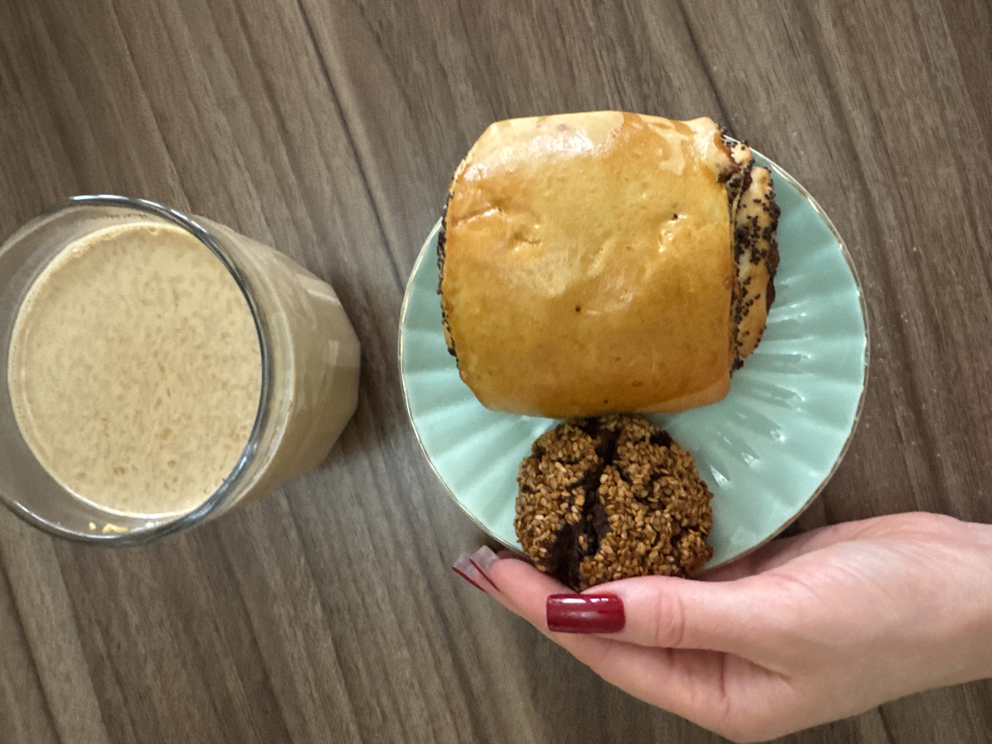 A high-angle medium shot shows a glass of milky-colored liquid, a pastry, and a sesame seed cookie on a light blue plate, being held by a hand with burgundy-colored fingernails. The background is a brown wood grain surface. The pastry is square, glazed, and has poppy seeds along one side. The scene is in an unknown location and city.