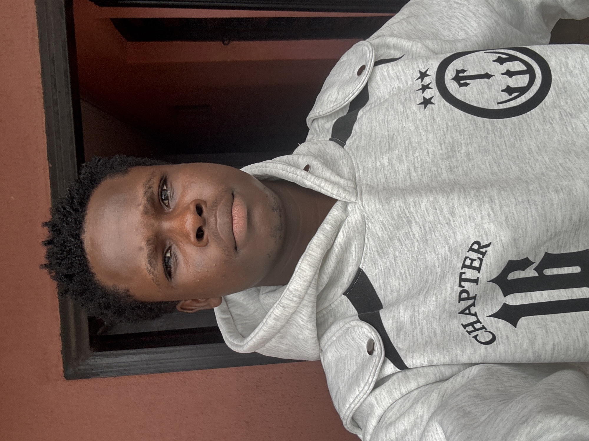 A young Black male, appearing to be in his late teens or early twenties, poses for a selfie in front of a dark doorway, likely in Ihiagwa, Nigeria. He has short, dark, curly hair and brown eyes. He is wearing a light gray hooded sweatshirt with black accents. The sweatshirt has the word "CHAPTER" and the number "16" printed in black on the front, along with a circular logo featuring three stars above a design that includes the letter "T" and trident-like elements. The background shows a reddish-brown wall and a dark interior, suggesting an indoor or covered outdoor setting. The lighting is somewhat dim, with a slight glare on his forehead.