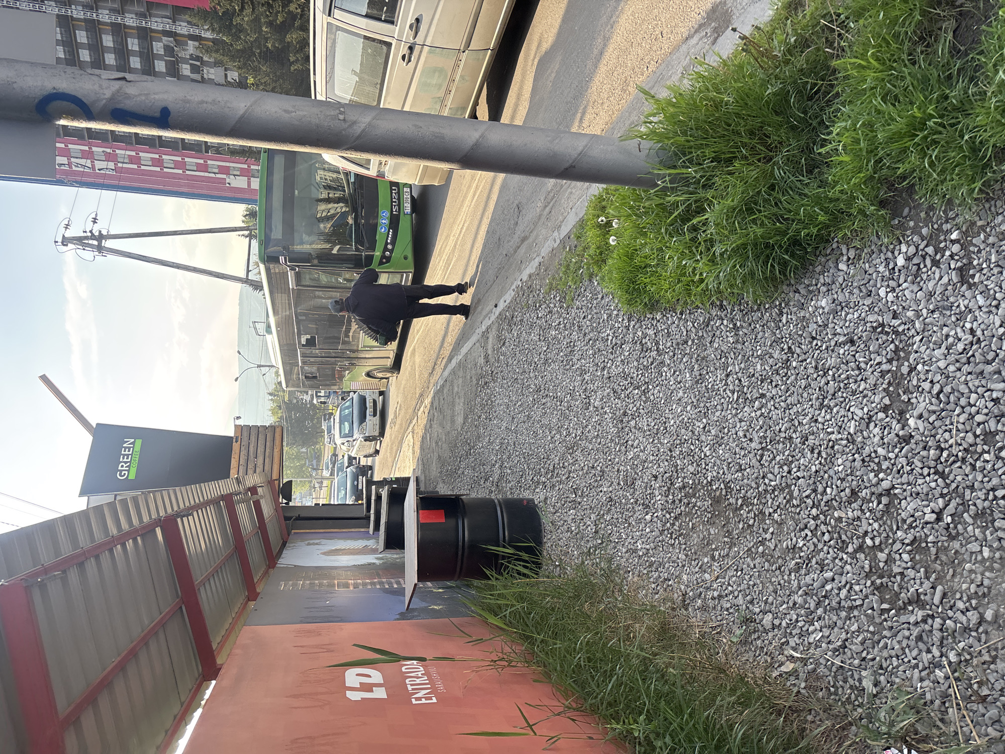 The image shows a street scene in Tbilisi, Georgia. A person is walking from right to left between a green bus and a parked minivan. The bus has a visible license plate. A utility pole stands near the sidewalk with grass and small white flowers. The foreground features a gravel-covered area, a black barrel, and a partially visible advertising structure with the text "1D Entrada" and "GREEN coffee." Buildings are visible in the background.