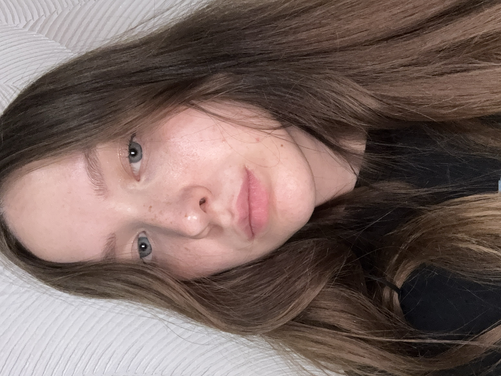 A close-up, horizontal portrait captures a young woman lying down and looking directly towards the viewer. Her head is tilted, giving a diagonal composition to the image. She has long, wavy medium-to-dark brown hair with subtle lighter highlights, which cascades around her head and frames her face. Her complexion is fair, with visible freckles scattered across her nose and upper cheeks. Her eyes are a clear blue-gray, with a visible catchlight in her left pupil, indicating a light source. Her lips are a natural, soft pink, slightly parted. She appears to be wearing a dark-colored top, possibly a t-shirt, which is partially visible behind her hair on the right side of the frame. The background consists of a textured, off-white surface, likely a pillow or mattress, suggesting an indoor setting such as a bedroom. The lighting is soft and even, highlighting her features and suggesting an intimate or relaxed atmosphere. Her expression is calm and neutral. No text is visible in the image. The person is located in Chaikovsky, Russia.