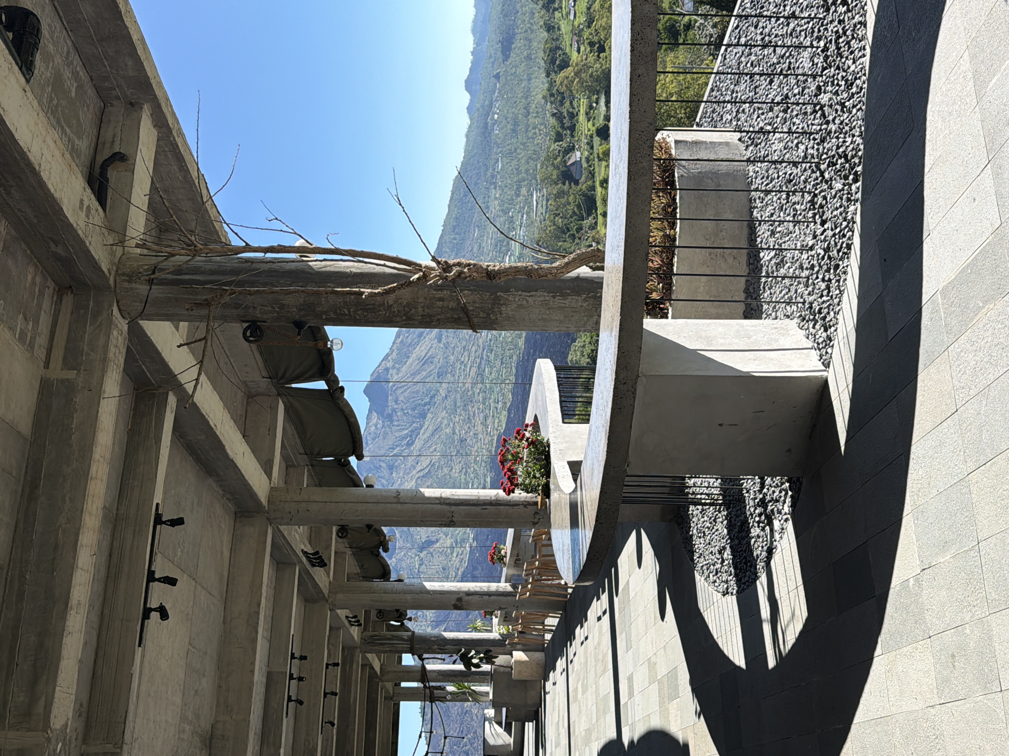 The image depicts an outdoor structure with a view of mountains in Kedisan, Indonesia. The structure features a concrete frame with round concrete posts, some of which support canopies and plant boxes containing flowers. There are small lights mounted on the concrete walls. Alongside the structure is a walkway covered with gray stones and patterned gray tile, casting shadows. A railing separates the walkway from a section covered in smaller gray pebbles. The view beyond the structure shows a mountainous landscape with vegetation under a clear blue sky.