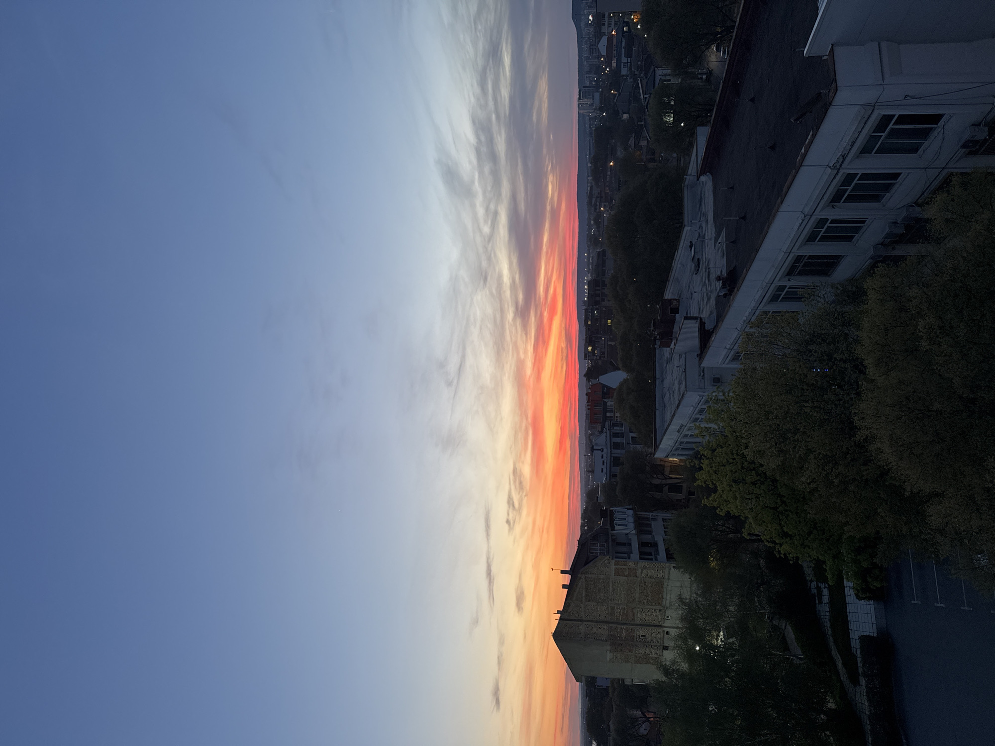 This image captures a panoramic view of Varna, Bulgaria, at either sunrise or sunset, taken from an elevated perspective. The sky dominates the left and central portions, displaying a dramatic gradient of colors. On the far left, the sky is a soft, clear blue, transitioning towards the center and right into vibrant hues of orange, red, and yellow, characteristic of dawn or dusk. Long, wispy clouds stretch across the sky, catching these brilliant colors.
Below the horizon line, a sprawling cityscape unfolds. Numerous buildings, likely residential and commercial, line the streets, their forms silhouetted against the bright sky and illuminated by emerging artificial lights in the distance. A faint outline of distant hills or mountains is visible on the horizon.
In the foreground and mid-ground, the rooftops and upper floors of closer buildings are prominent. These structures feature multiple windows, some with visible air conditioning units, and dark, flat roofs. Lush green trees, some densely foliated, are interspersed among the buildings, adding a natural element to the urban landscape. A parking lot with white markings can be seen in one of the darker foreground sections. The overall scene is peaceful and highlights the natural beauty of the sky contrasted with the urban environment. There is no visible text in the image.