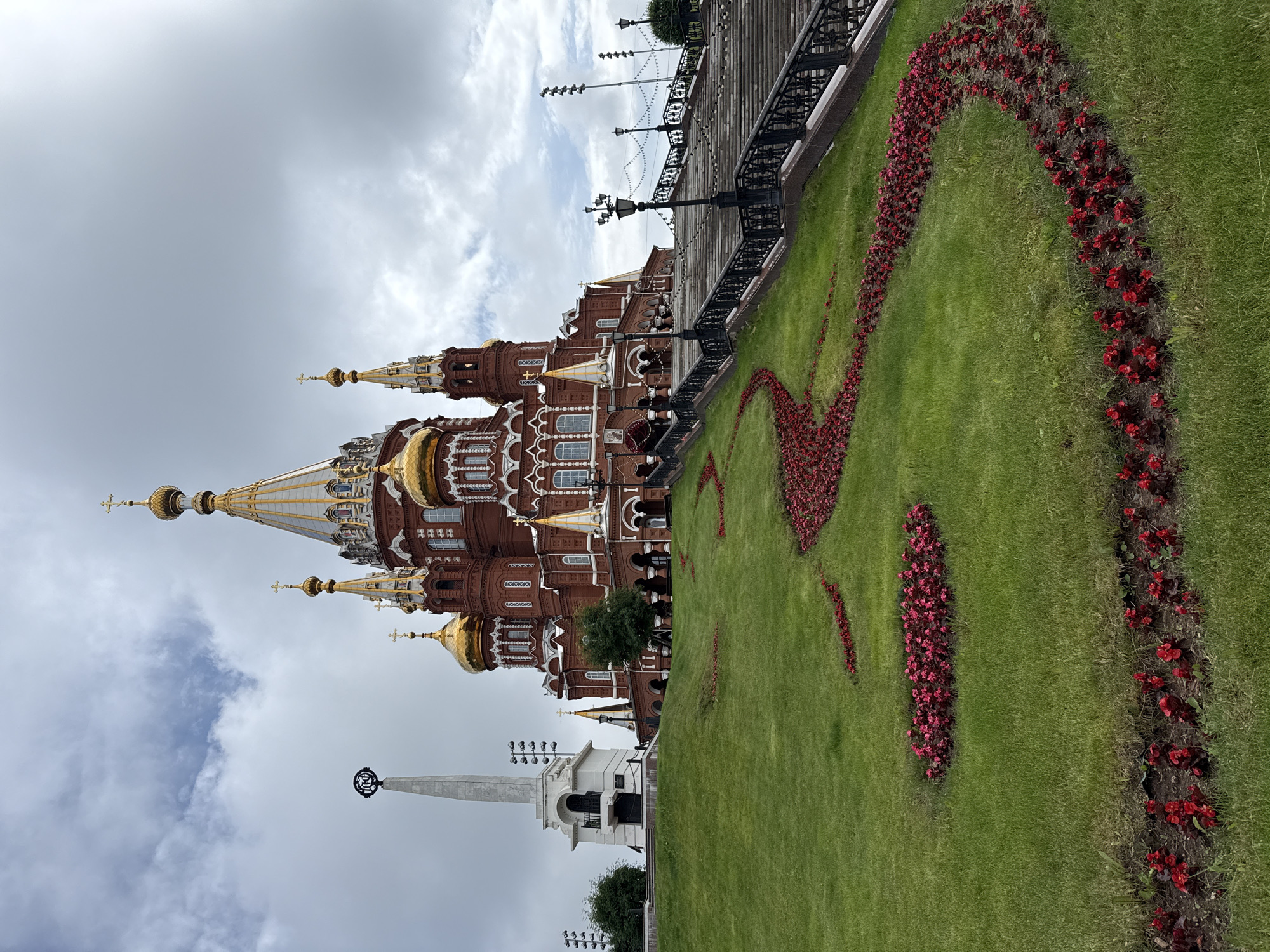 The image depicts the Holy Trinity Cathedral in Izhevsk, Russia, under a cloudy sky. The grand red brick cathedral features golden domes and a central spire, with smaller spires surrounding it. In the foreground, a lush green lawn is adorned with a serpentine flower bed of vibrant red flowers. A wide stone staircase with black ornate railings leads up to the cathedral's entrance. To the left of the cathedral, a tall white obelisk with a circular emblem at its apex stands on a raised platform, with a monument base below it. The scene is set on a hill, offering a panoramic view of the cathedral and its surroundings. No people are visible in the image.