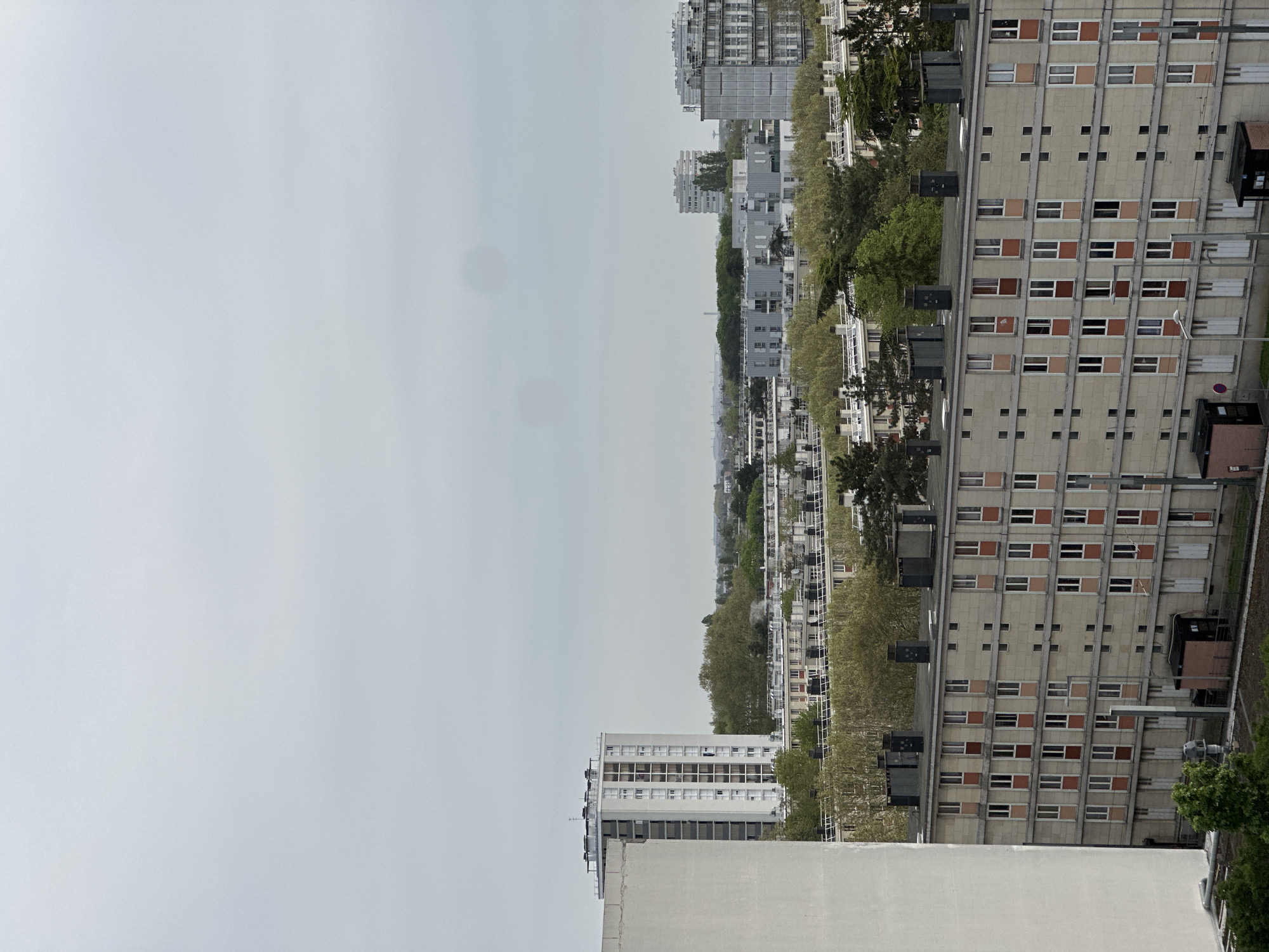 The image shows a cityscape of Sarcelles, France, under a cloudy sky. The architecture is characterized by numerous apartment buildings, many of which have a modular, grid-like facade with rows of windows. The color palette is muted, dominated by gray, beige, and brown tones. Trees are interspersed between the buildings, with bare branches indicating a transition to spring. A tall structure is seen in the distance. There are no people visible. The setting appears to be a residential area within the city.