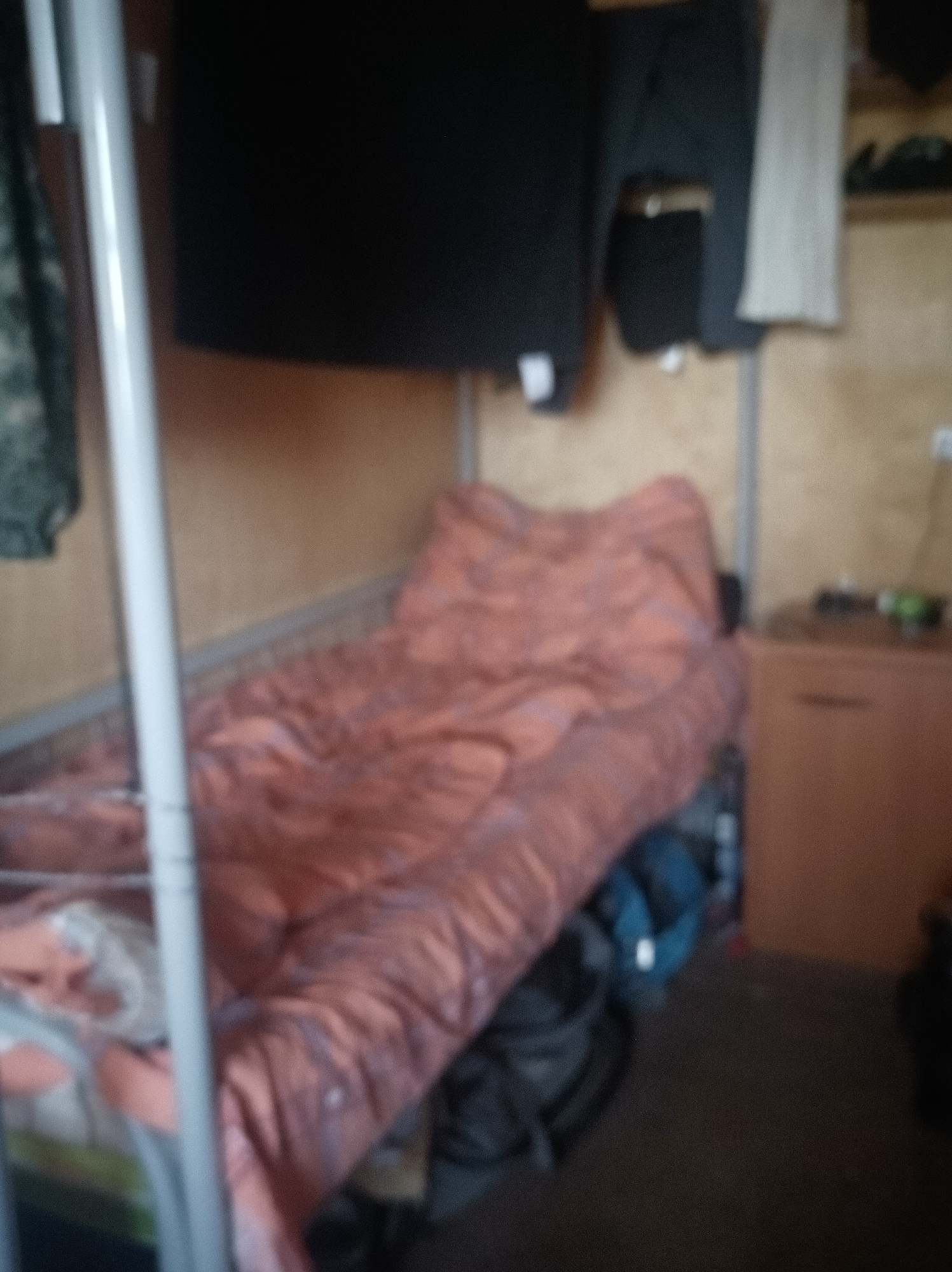 The image depicts a dimly lit, cramped living space, likely a dormitory room or a small dwelling in Vybenka, Russia. The focal point is a narrow bed with a salmon-colored comforter, appearing somewhat disheveled. To the left of the bed, a white metal frame of what appears to be a bunk bed is visible. Above the bed, dark clothing, possibly a sweatshirt and pants, is hung on a rack. A pale grey garment is also hanging nearby.
On the right side of the image, a wooden cabinet with a drawer is situated. On top of the cabinet, a small green object, possibly a bottle or container, is visible. The walls of the room are a light brown, wooden paneling. Various bags and dark objects are scattered on the floor beneath and to the side of the bed, suggesting a lived-in, possibly untidy environment. The overall impression is of a functional, but spartan, living quarters.