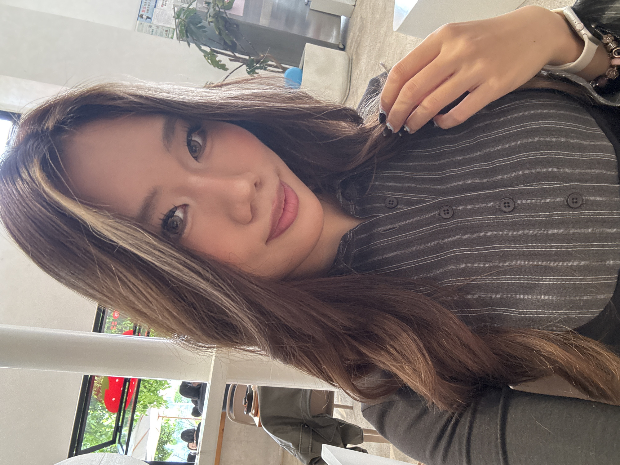 A woman with light brown hair, a streak of blonde in the front, brown eyes, and pink lips is looking at the camera. She is wearing a gray, pinstriped button-down shirt. She has a white smartwatch on her wrist, as well as a bracelet. Her right hand is near her face. The background is blurred, but it appears to be an indoor space, likely a cafe or restaurant. A plant and a white cabinet are visible. There are other people at tables in the background. A white pole is visible near a window. Based on visual clues, it appears this photo may have been taken in 江北城街道, China.