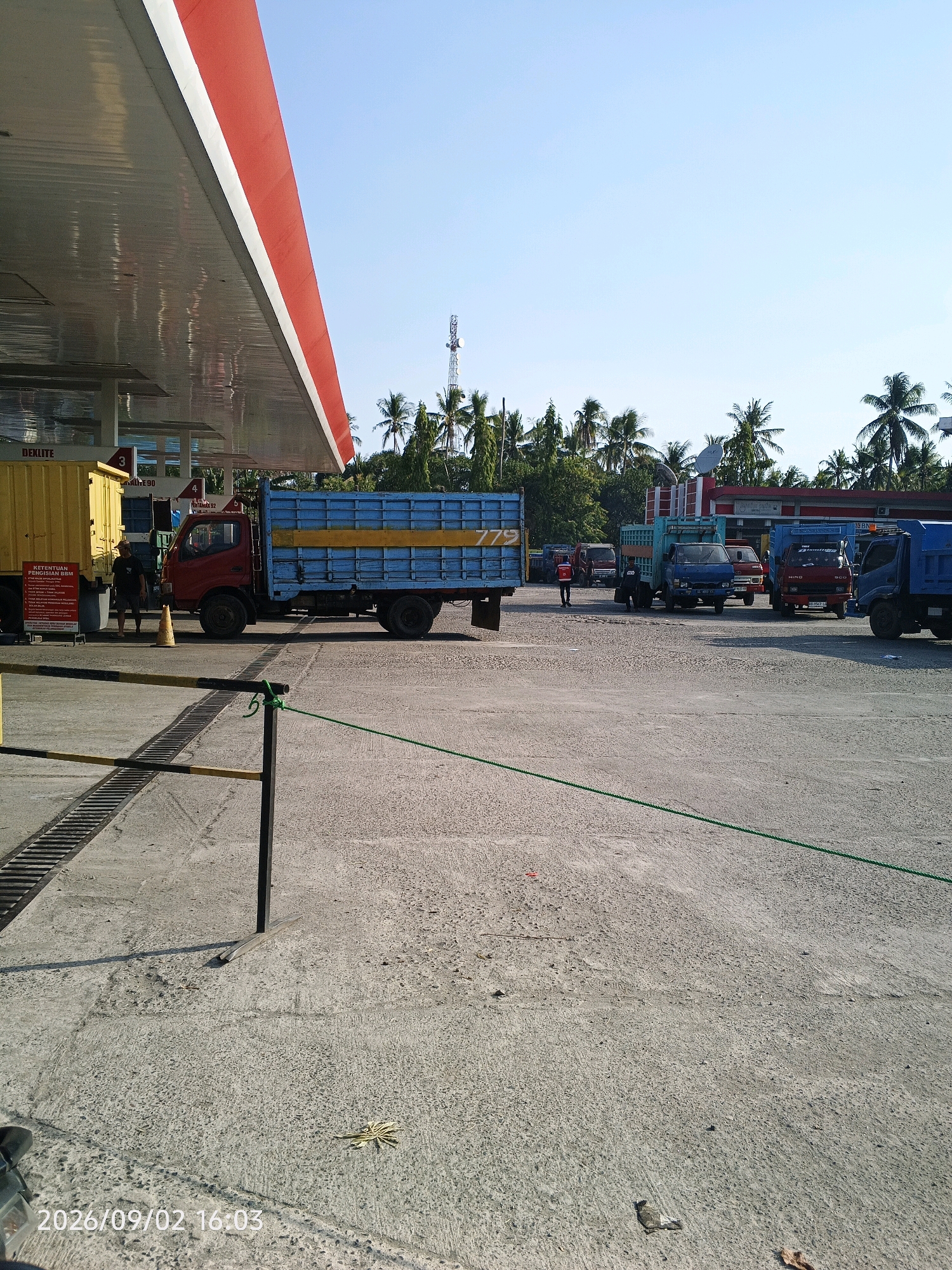 An outdoor scene captures a gas station in Pinrang, Indonesia, under a clear blue sky. The foreground is dominated by a concrete surface with a yellow and black striped barrier and a green rope tied to a black post. To the left, the roof of a gas station canopy extends into the frame, featuring red and white trim. A yellow truck is parked to the left of the canopy. In the middle ground, a red truck with a blue cargo bed displaying the number "779" is positioned. Several other trucks, including blue and red ones, are visible in the background, along with palm trees and a radio tower. A person in a red shirt is standing near the blue-cargo truck, and another person in dark clothing is walking past the yellow truck. The overall atmosphere suggests a normal, active day at the gas station.