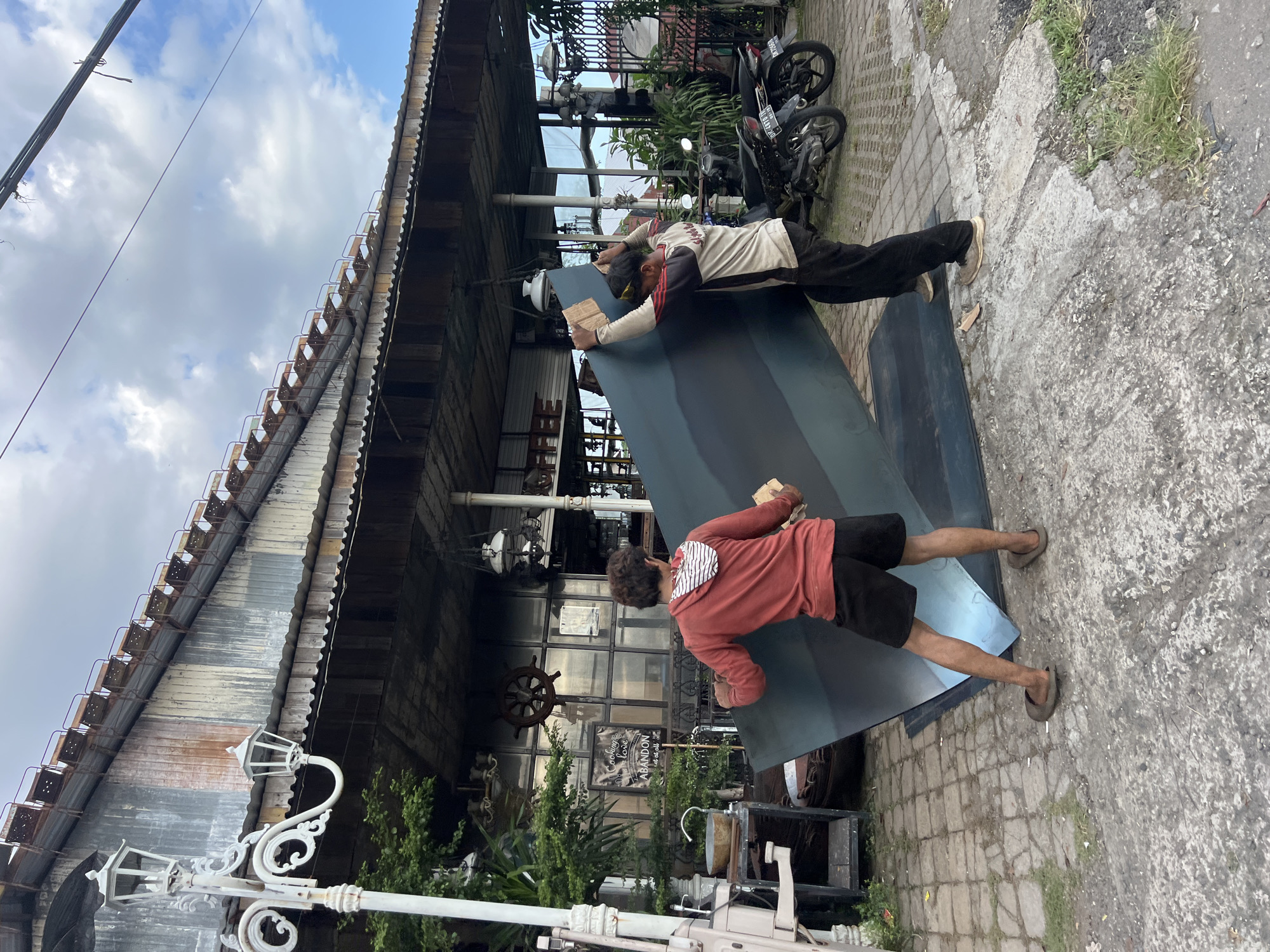 Two men are working on a large metal sheet outside a building in North Kuta, Indonesia. The man on the left wears a light-colored long-sleeved shirt, dark pants, and yellow-tinted goggles, and uses a wooden block to work on the sheet. The man on the right, facing away from the camera, wears a red sweater and black shorts. The building behind them has a rustic, industrial aesthetic with metal siding and a weathered roof. Details such as a ship's wheel, hanging lights, and plants are visible in front of the building. Two motorcycles are parked nearby. The area appears to be a street or sidewalk paved with stone blocks. The sky is cloudy.