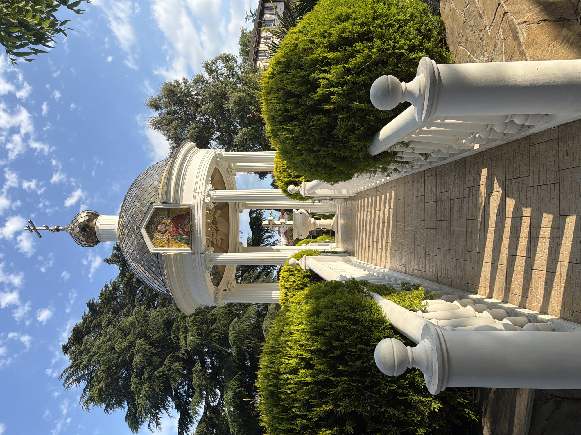 The image depicts a sunlit outdoor scene with a focus on a white architectural structure with a domed roof, reminiscent of a chapel or a gazebo, surrounded by lush green trees and manicured bushes. The structure features ornate white pillars and a decorative railing. A tiled pathway leads towards the structure, casting long shadows indicative of the time of day. The sky is a bright blue with scattered white clouds. The overall impression is one of peace and serenity, possibly a religious or contemplative space. The time of day appears to be late morning or early afternoon, given the angle of the sun and the shadows. No people are visible in the frame. The location context of Sochi, Russia, suggests a potentially temperate climate. The architecture hints at a style that might be found in Eastern Europe or countries with similar cultural influences.