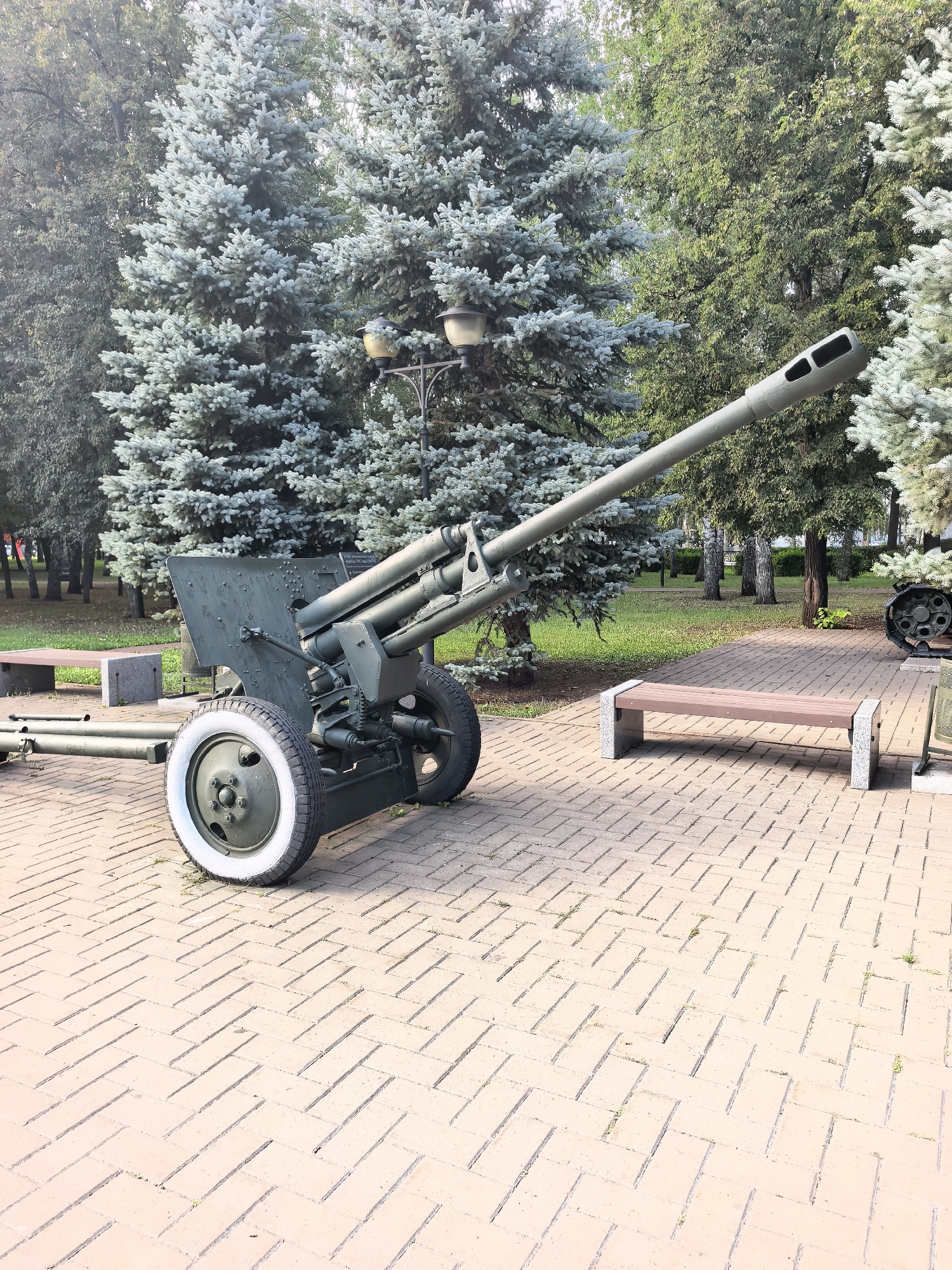 The image shows a historical artillery piece, likely a towed gun, displayed outdoors on a paved walkway in Sterlitamak, Russia. The gun is painted olive drab and features two large spoked wheels with white tires. It is positioned on a brick-patterned walkway, with a wooden bench with concrete supports visible to the right. Behind the gun, a stand of mature evergreen trees, possibly blue spruce, creates a lush backdrop. A lamppost with two lights is also visible among the trees. The scene appears to be part of a park or memorial site, given the presence of the historical military hardware and seating. No people are present in the image.
