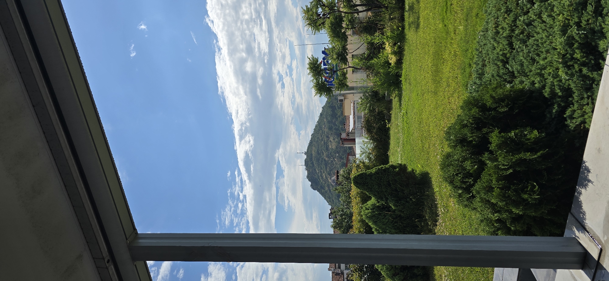 The image presents an outdoor scene in Plovdiv, Bulgaria, viewed from a partially covered structure, likely a balcony or terrace, under clear daytime conditions with scattered white cumulus clouds in a blue sky. The top of the frame features the underside of a light grey ceiling or beam, exhibiting small water droplets along its horizontal edge. A prominent light grey vertical support column is visible on the left side, extending from the top towards the bottom of the frame. Below the column and extending across the bottom, a grey paved surface is visible.
The midground is dominated by a meticulously maintained green lawn, interspersed with various sculpted evergreen bushes and shrubs. Beyond the garden space, residential buildings with traditional terracotta-tiled roofs are visible. On the right, a multi-story building features a large, partially obscured blue and red sign. The background is anchored by a large, densely vegetated hill, specifically Sahat Tepe (Danov Hill), one of Plovdiv's distinct "tepes," which is topped with multiple communication towers and antennas. No human subjects, animals, or explicit activities are observed within the frame.