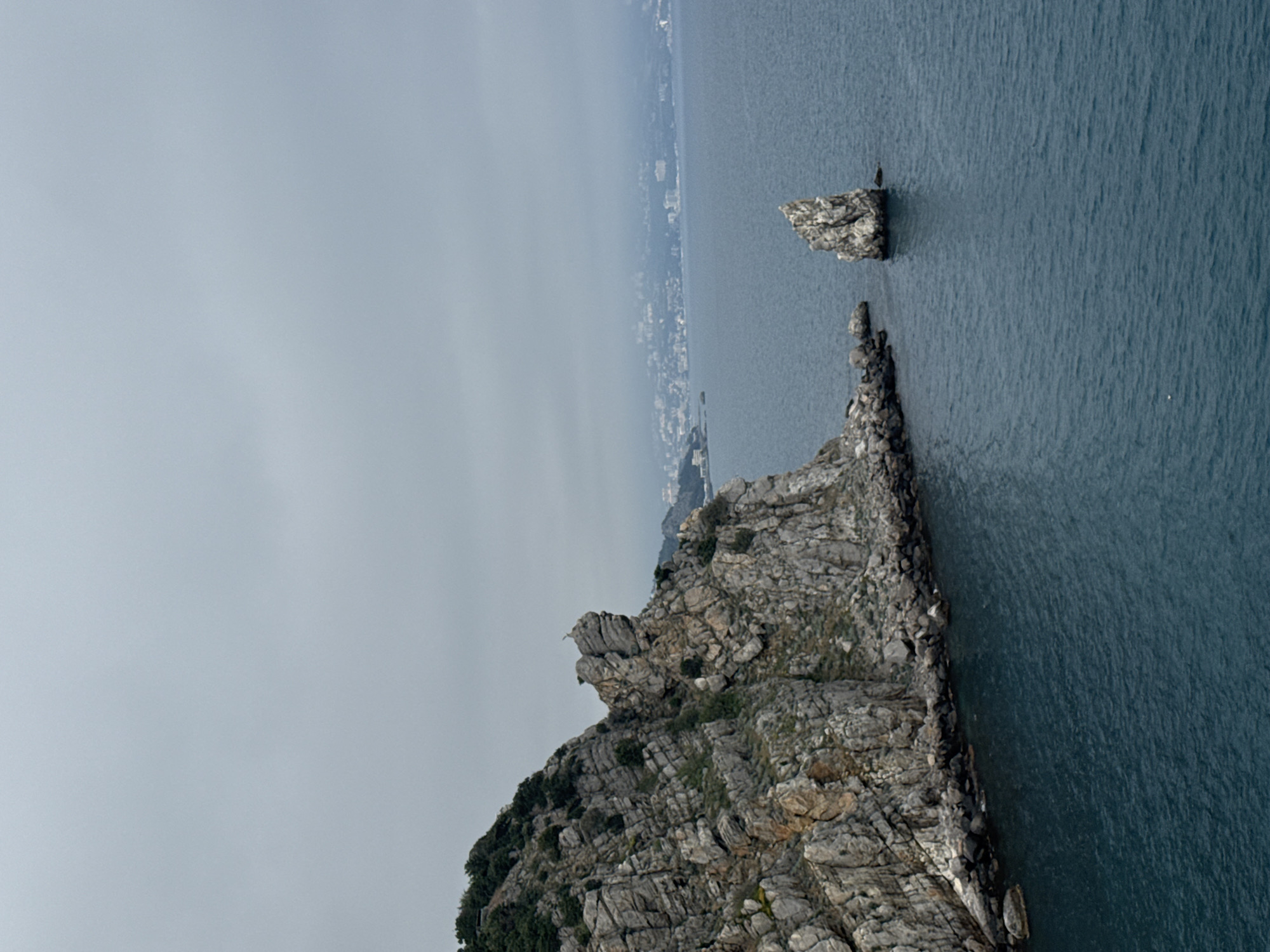 A landscape-oriented image shows a rocky coastline meeting the sea. The frame is dominated by a large, craggy cliff face with a rough texture, covered in patches of greenery. The cliff extends into the water, forming a jagged edge where land meets the sea. Further offshore, there is a smaller, isolated rock formation standing in the water. The sea is a dark shade of blue and has a rippled surface, suggesting a slight breeze. In the distance, the vague outline of a coastal city can be seen under an overcast sky, placing the image in the city of Гаспринский поселковый совет, Ukraine. The sky is a flat, gray color, indicating either a cloudy day or a time of day with soft lighting.